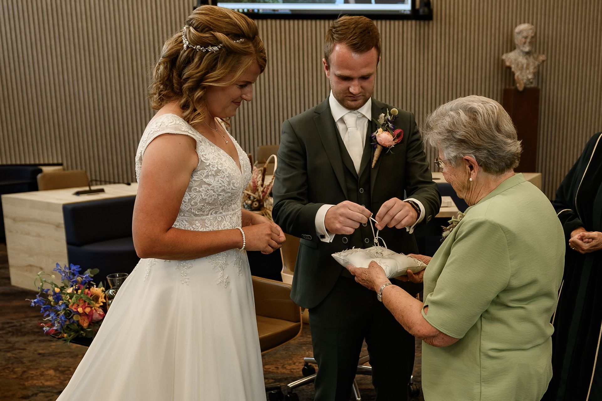 Bruid en bruidegom wisselen ringen uit tijdens een ceremonie. Een oudere vrouw houdt het ringkussen vast.