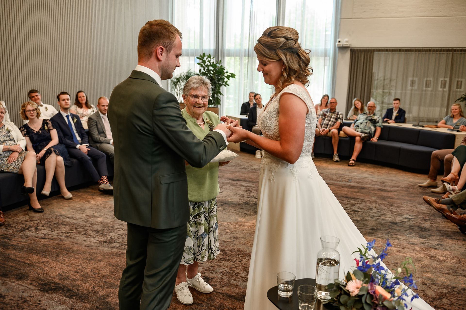 Een echtpaar wisselt ringen uit tijdens een huwelijksceremonie binnenshuis, die wordt voltrokken door een oudere vrouw. Gasten zitten.