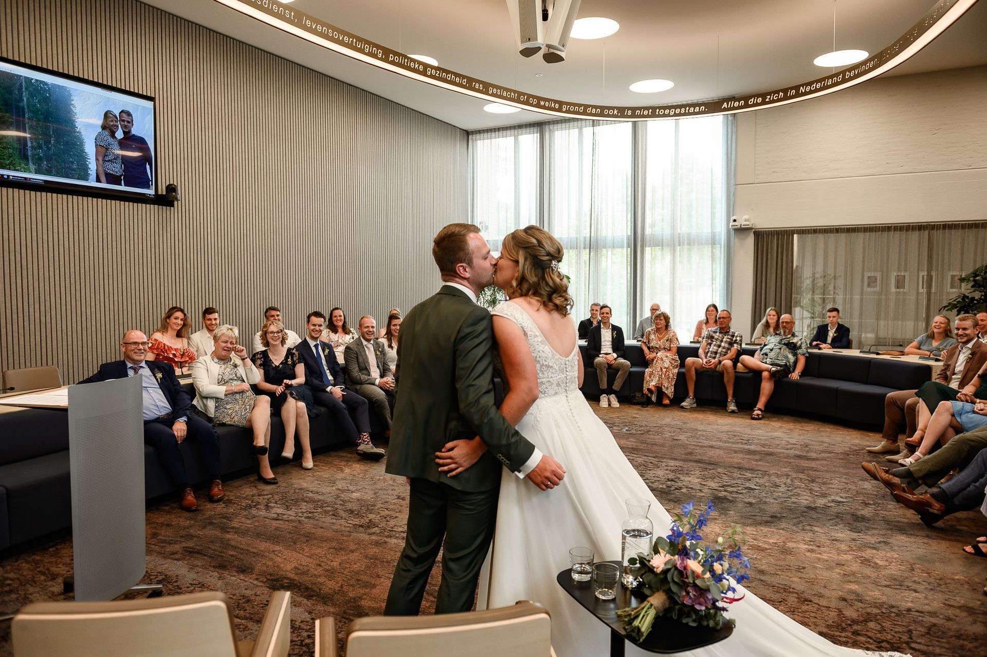 Een bruidspaar kust elkaar na de huwelijksceremonie, omringd door gasten in een moderne zaal met een groot raam.