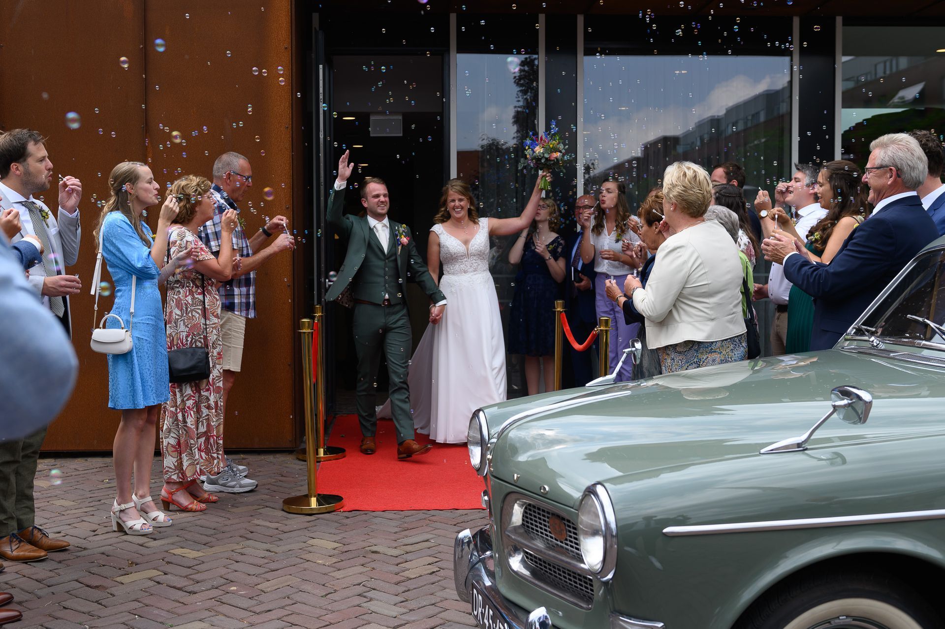 Het pasgetrouwde stel verlaat de locatie onder begeleiding van champagne en applaus; een oldtimer is op de voorgrond te zien.