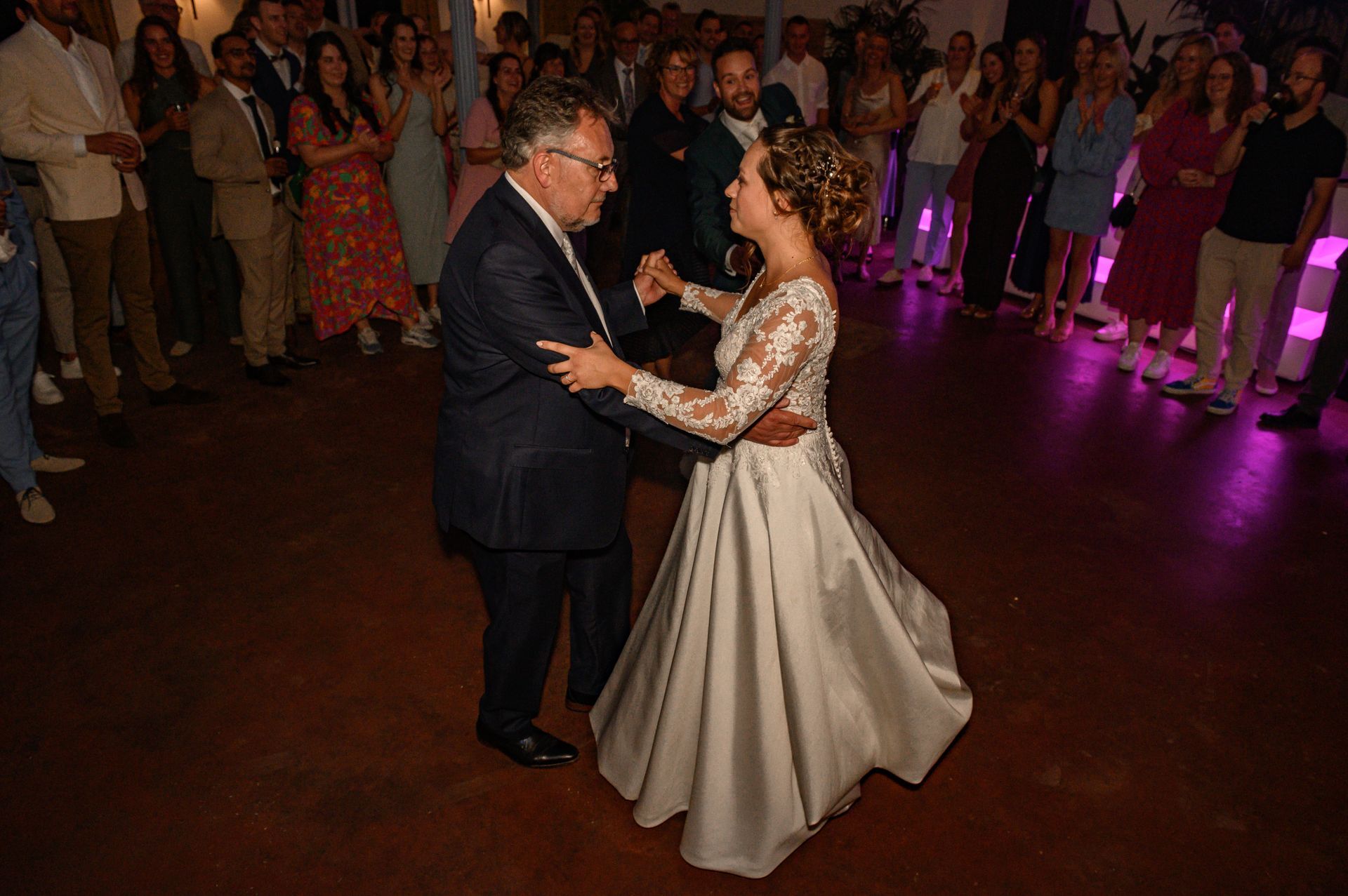 De bruid danst met een man, waarschijnlijk haar vader, op een huwelijksreceptie. Gasten kijken toe vanaf de zijlijn.