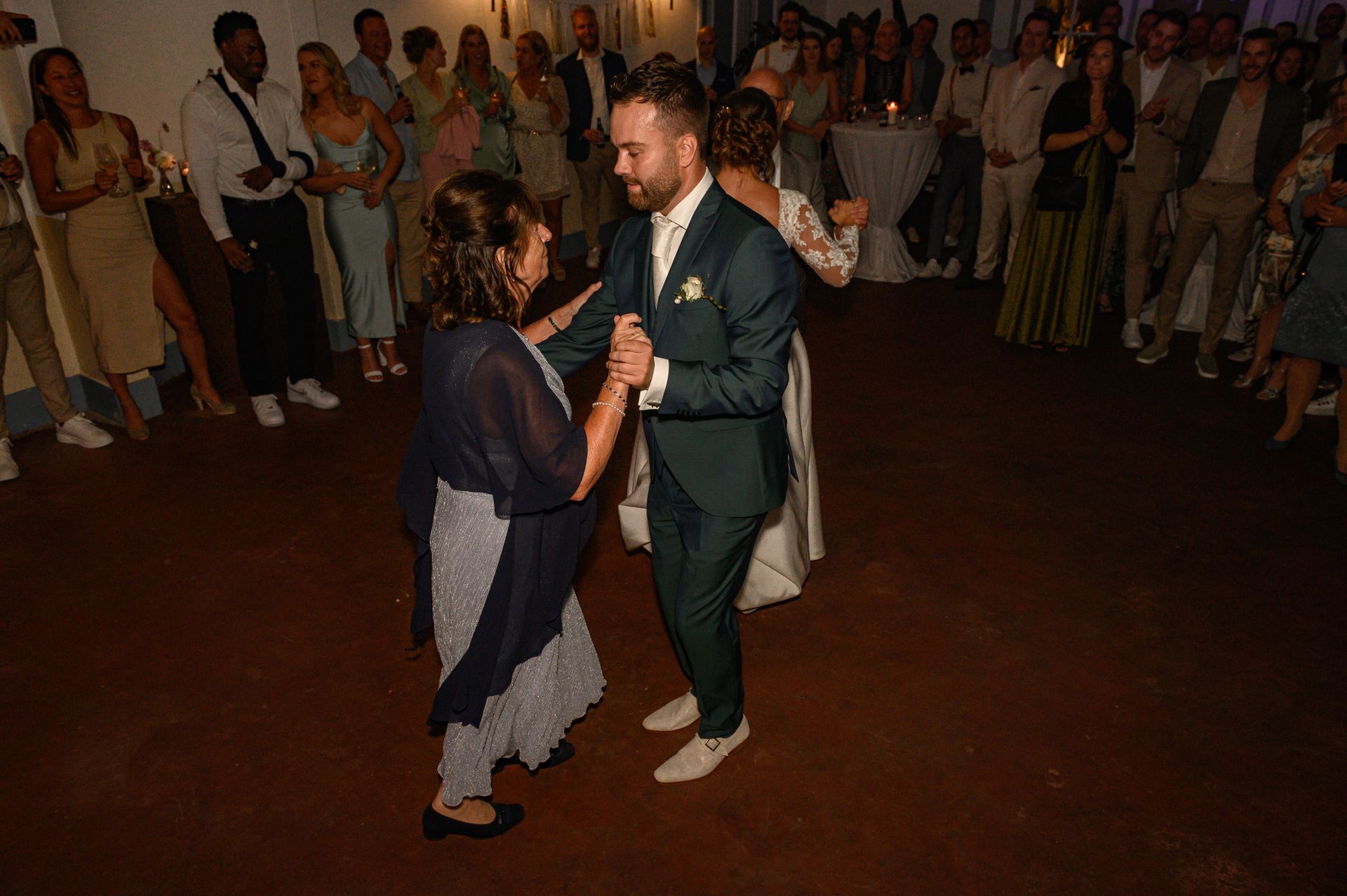 De bruidegom danst met een vrouw op een houten vloer tijdens een huwelijksreceptie. Gasten kijken op de achtergrond toe.