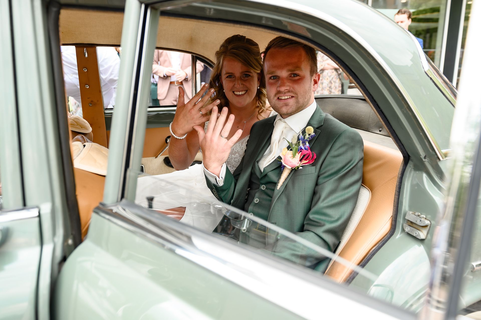 Pasgetrouwd stel in een vintage groene auto, lachend en hun ringen tonend.