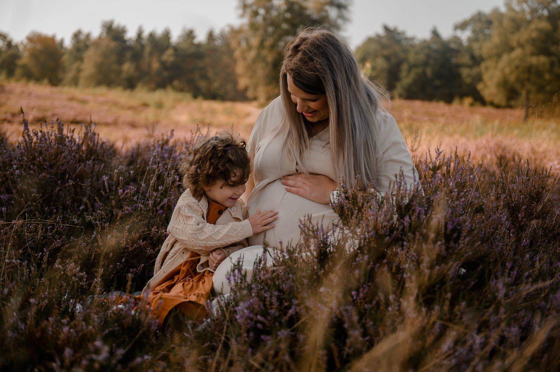 Een zwangere vrouw zit met een kind tussen paarse bloemen in een veld in de buitenlucht. Beiden raken de buik van de zwangere vrouw aan.
