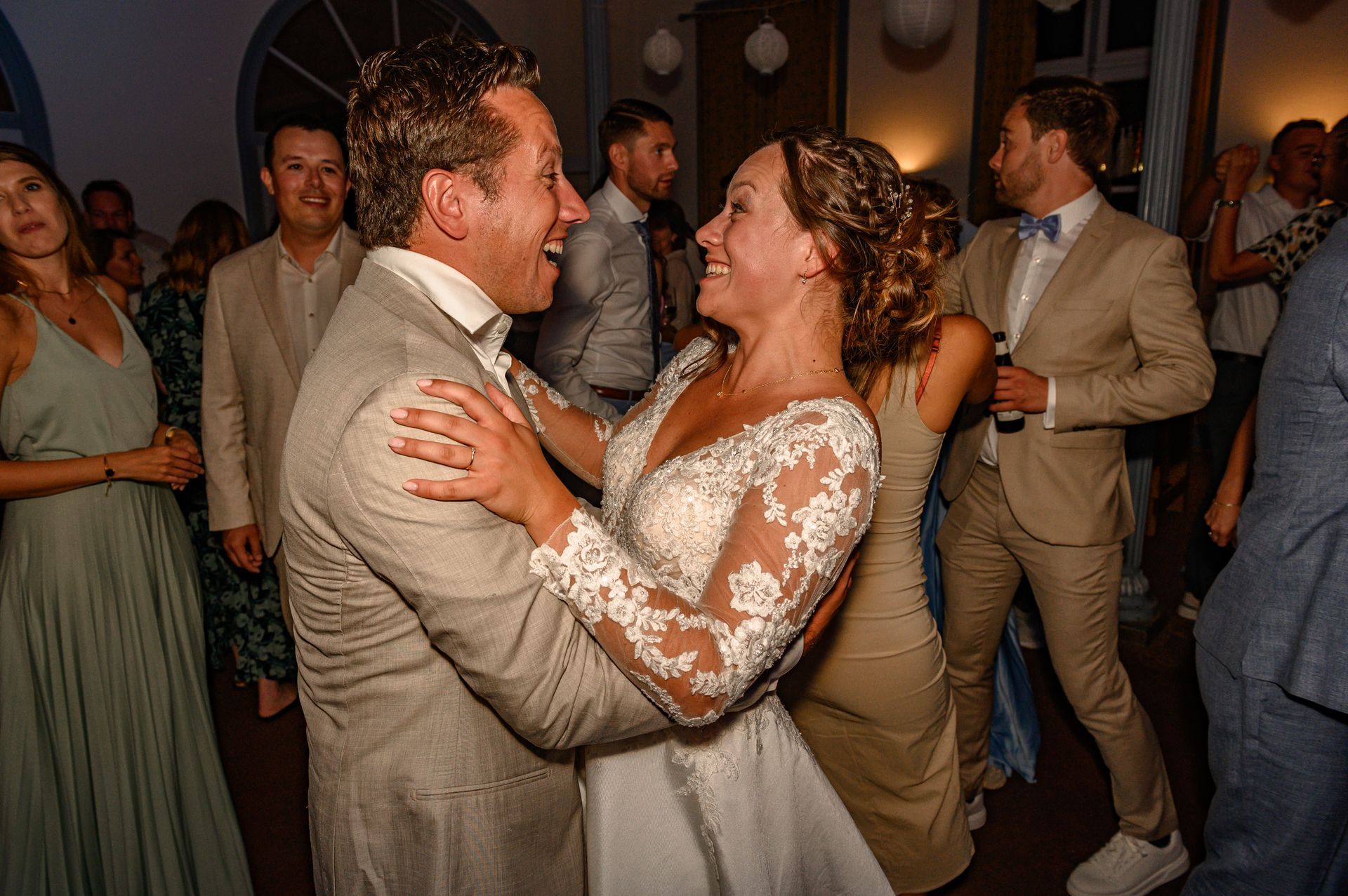 Bruid en bruidegom dansen op de huwelijksreceptie, stralend en lachend. Gasten op de achtergrond.