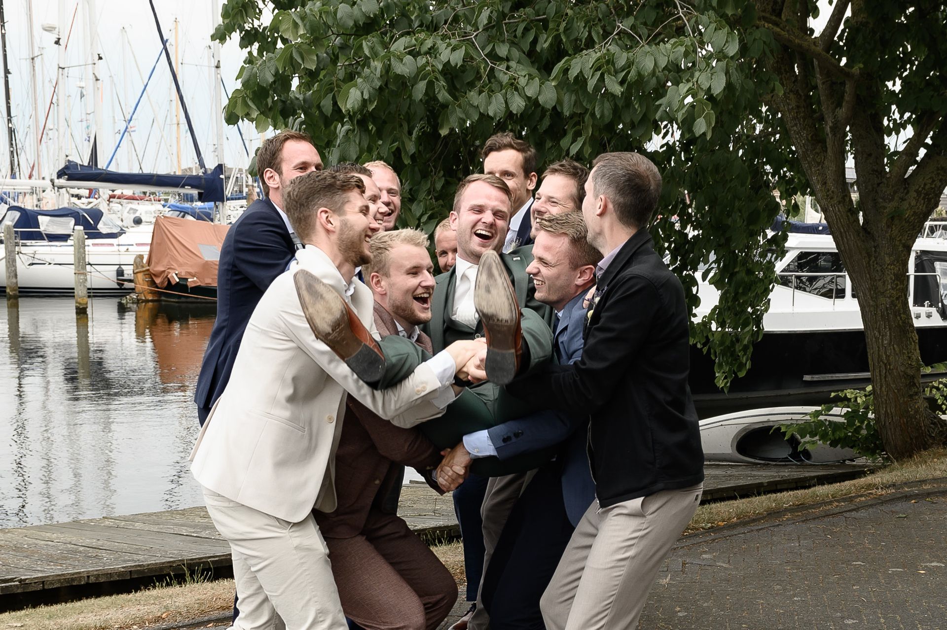 Een groep mannen in pakken staat lachend dicht bij elkaar bij een jachthaven, feestvierend.