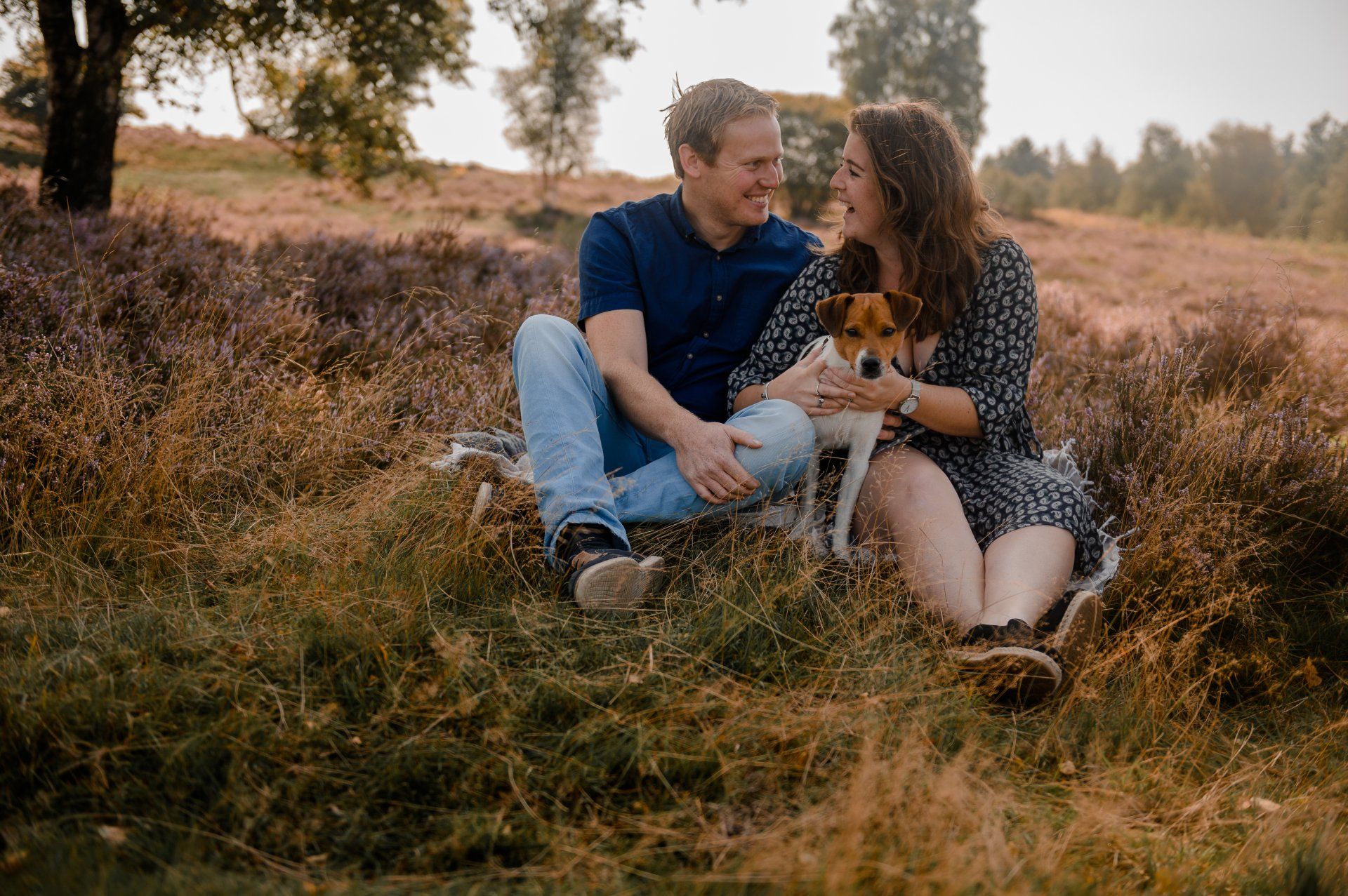 Een stel zit op een deken in een veld met paarse bloemen, aait een hond en lacht.