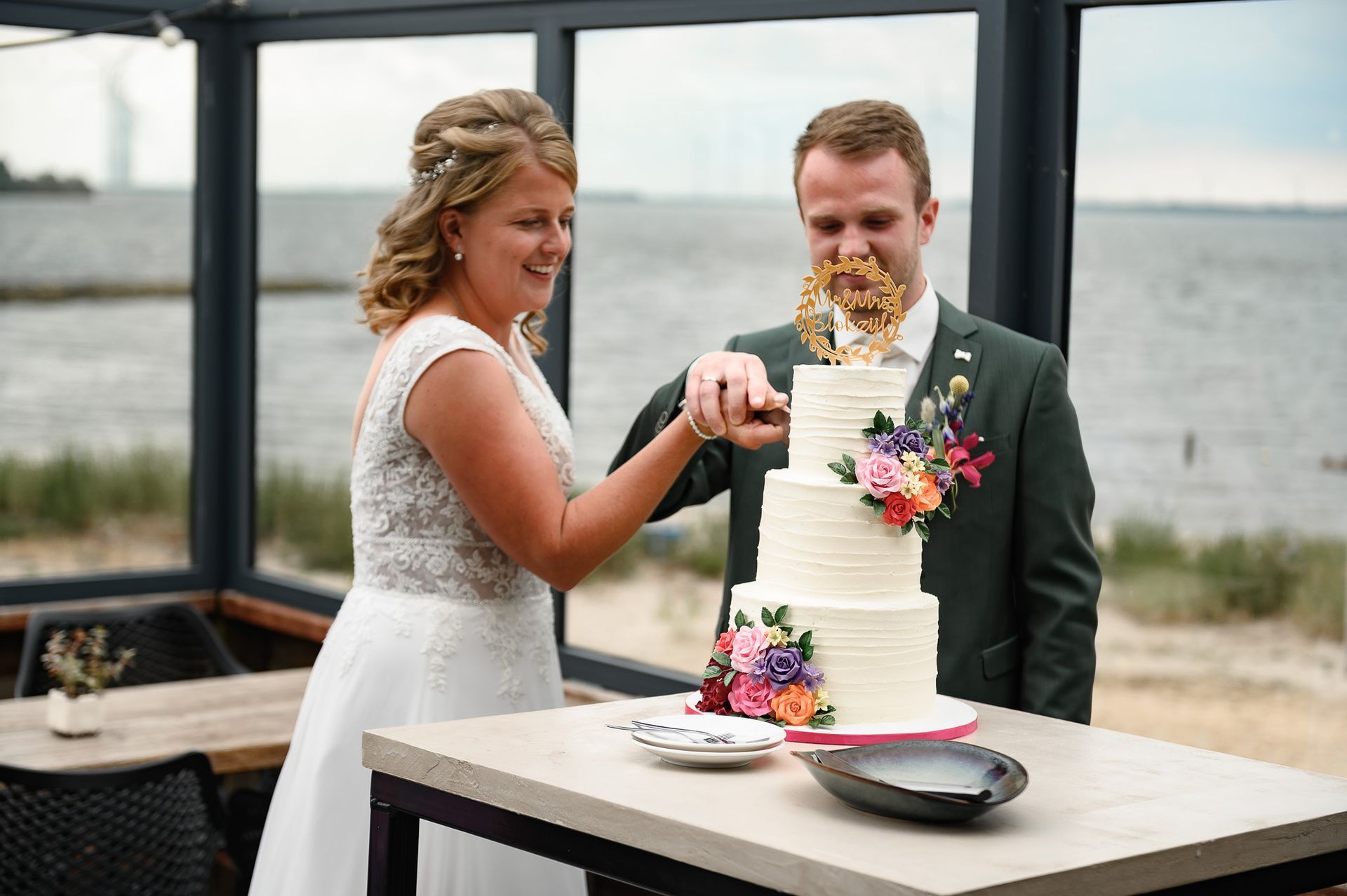 Bruid en bruidegom snijden een bruidstaart met meerdere lagen en bloemversieringen aan, met uitzicht op zee op de achtergrond.