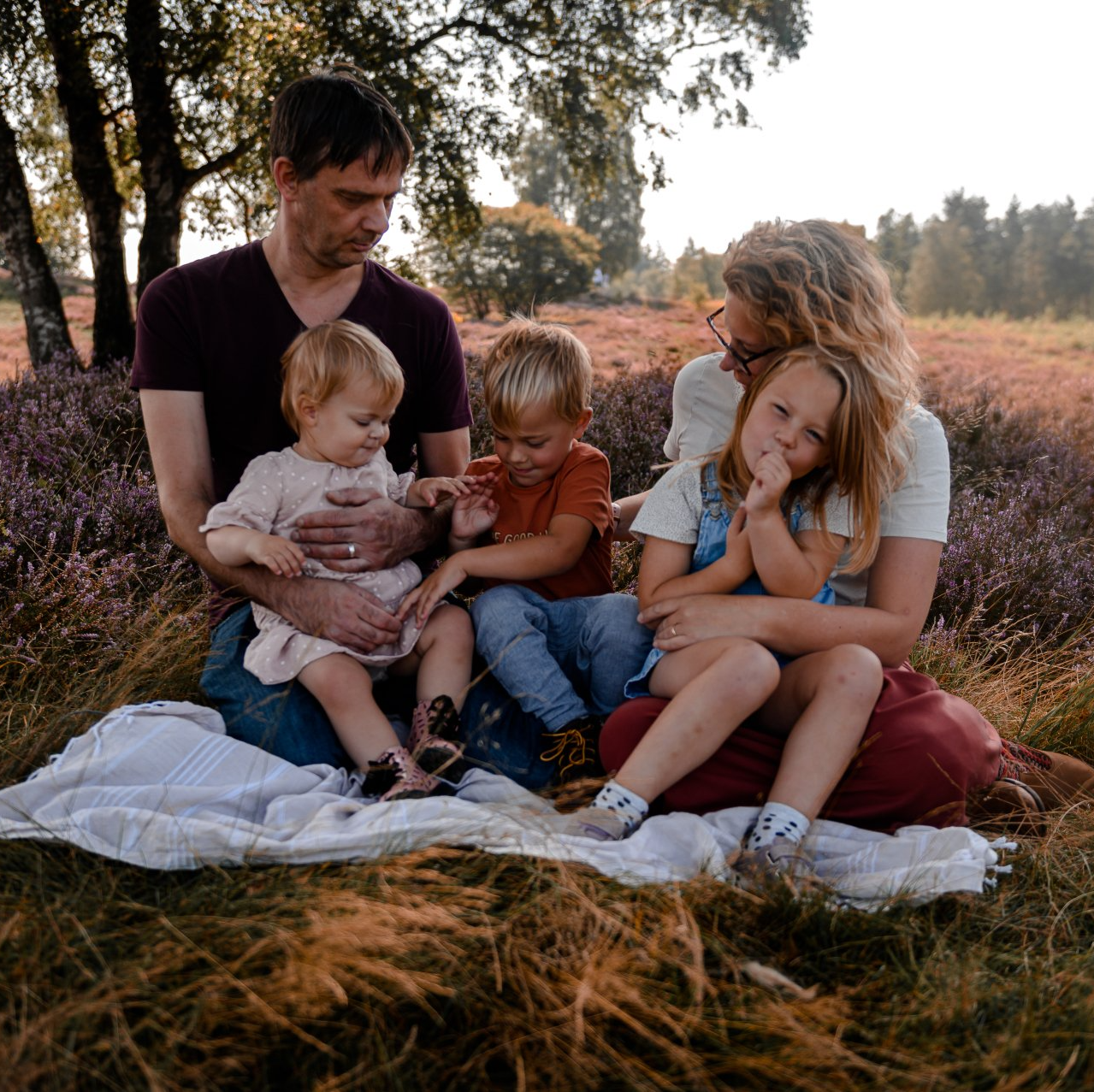 Een gezin zit op een deken in een veld met paarse bloemen; de kinderen kijken naar de camera.