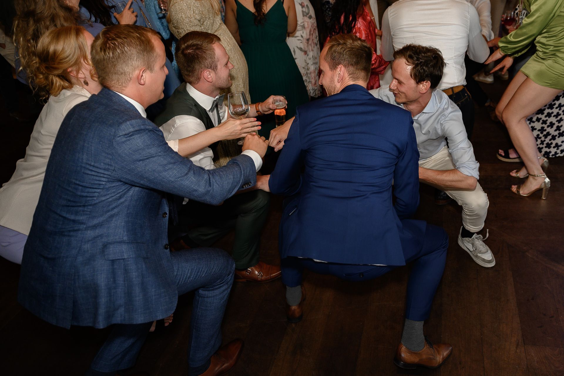 Mensen dansen en zitten gehurkt op een houten vloer, sommigen in pak, tijdens een levendig evenement.