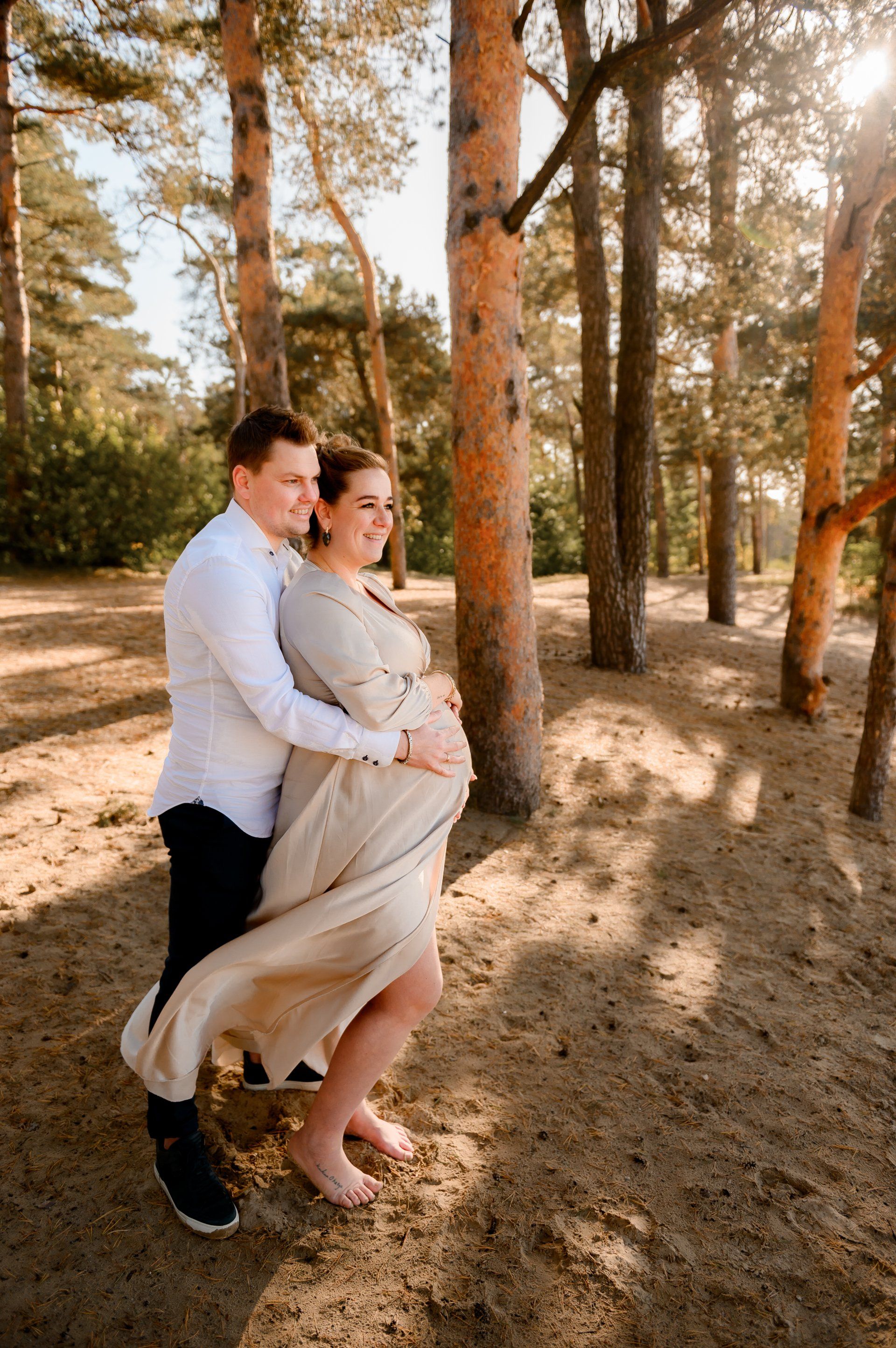 Een stel in een bos, de vrouw is zwanger. De man omhelst haar van achteren, beiden kijken in de verte. Lichtbruine jurk, zandkleur.