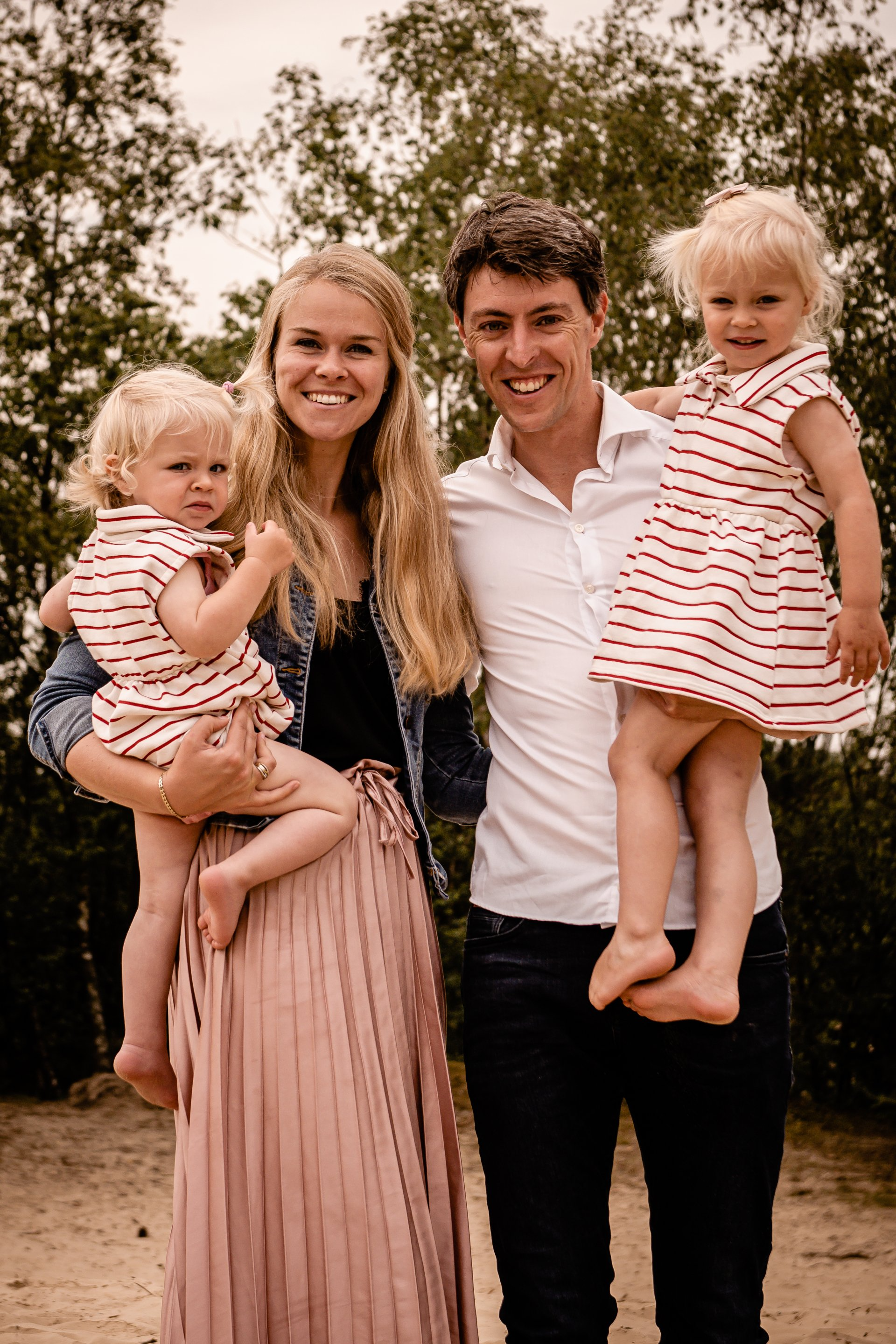 Een gezin van vier lacht en poseert buiten: twee jonge kinderen worden door hun ouders vastgehouden.