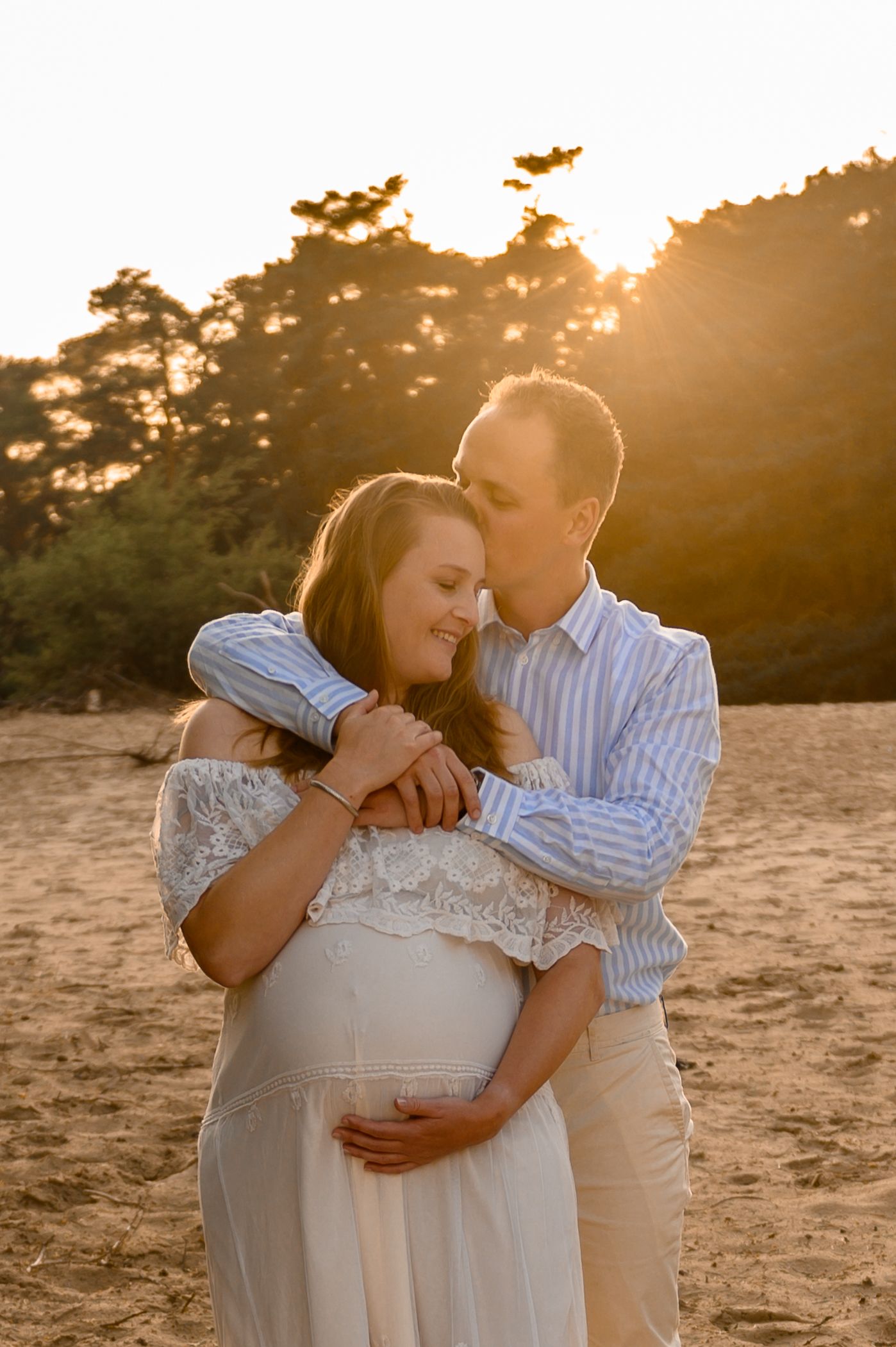 Een stel omhelst elkaar op het strand; de man kust de vrouw op haar voorhoofd, zij houdt haar zwangere buik vast. Zonsondergang op de achtergrond.