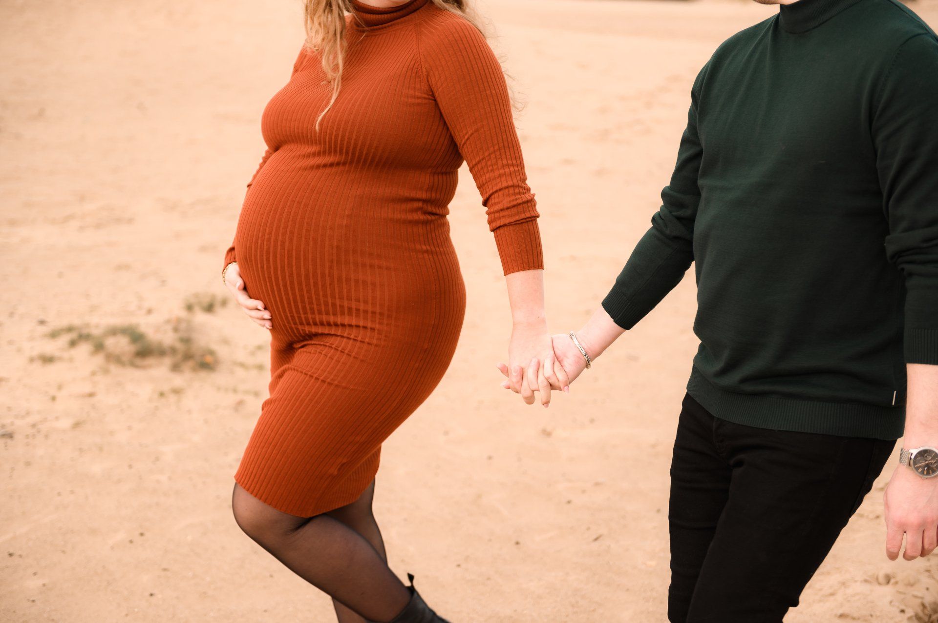 Een zwangere vrouw in een roestbruine jurk loopt hand in hand met een persoon in een groene trui, terwijl ze buiten wandelt.