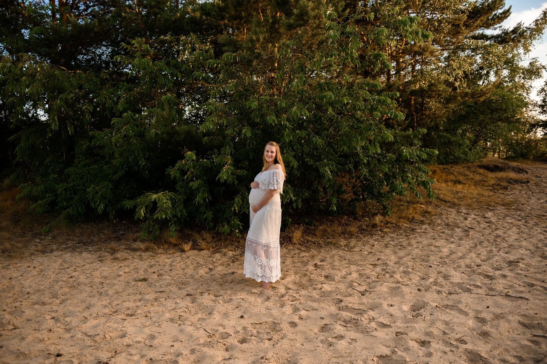 Een zwangere vrouw in een witte kanten jurk staat op een zandstrand voor een groene struik.