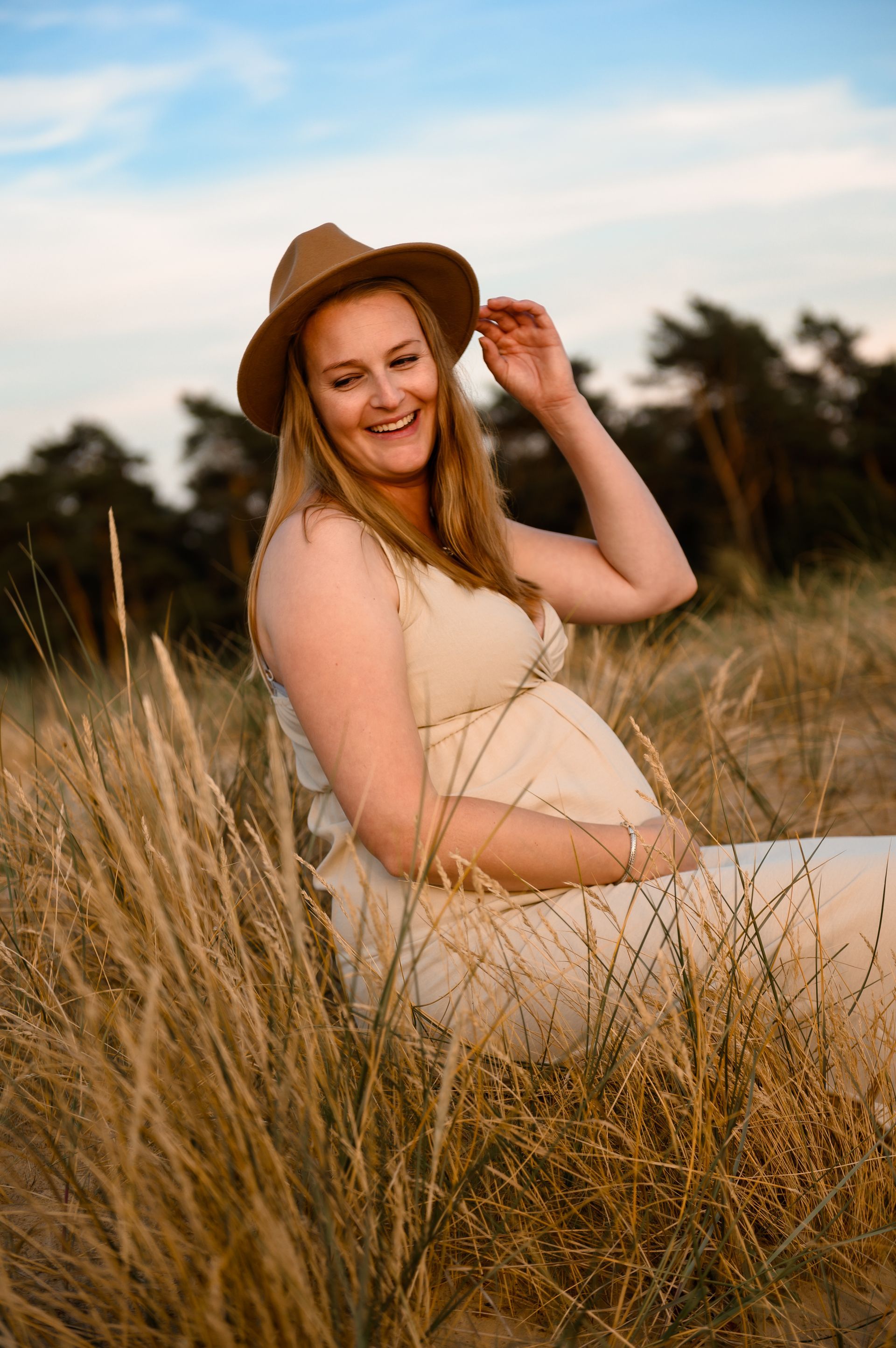 Een zwangere vrouw met een hoed op, lachend, zit in het hoge gras en legt haar hand op haar buik.