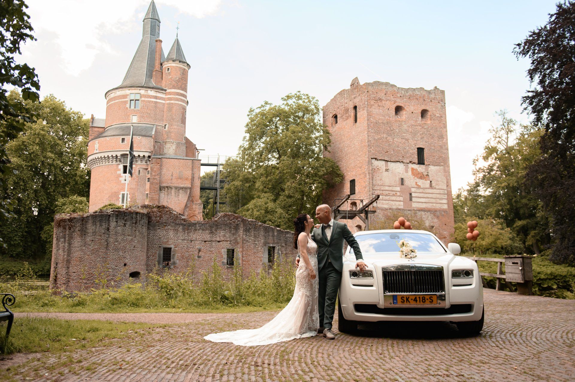 Een stel poseert naast een witte Rolls Royce voor een kasteel. De bruid draagt ​​een trouwjurk, de bruidegom een ​​pak.