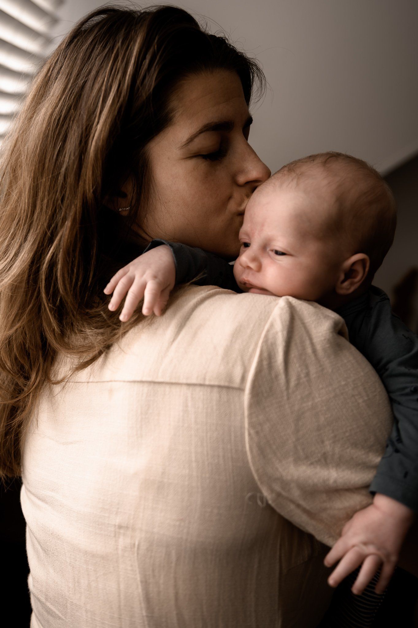Vrouw kust een baby op het hoofdje; de ​​baby rust op haar schouder. Warm licht valt door een raam.