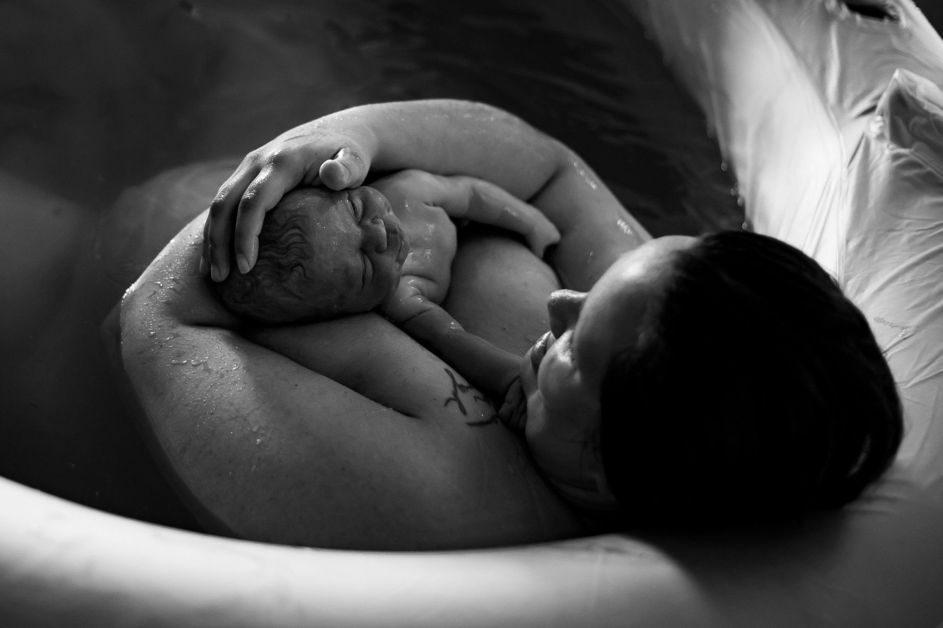 Een vrouw wiegt een pasgeboren baby in een bevalbad, haar gezicht dicht bij de baby, zwart-wit.