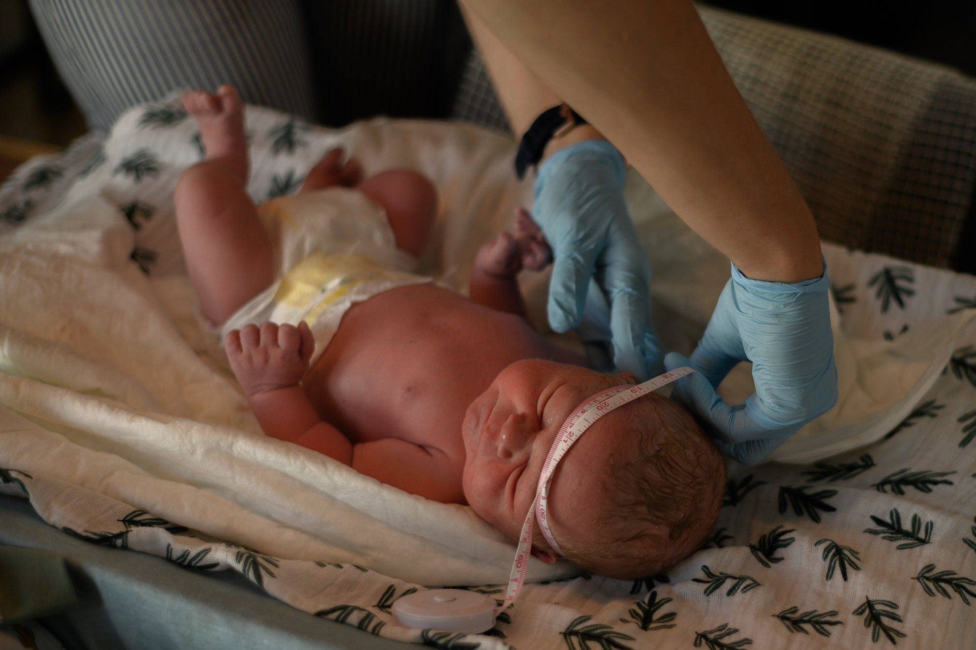 Een persoon met blauwe handschoenen meet het hoofdje van een pasgeboren baby met een meetlint. De baby ligt op een deken met een patroon.