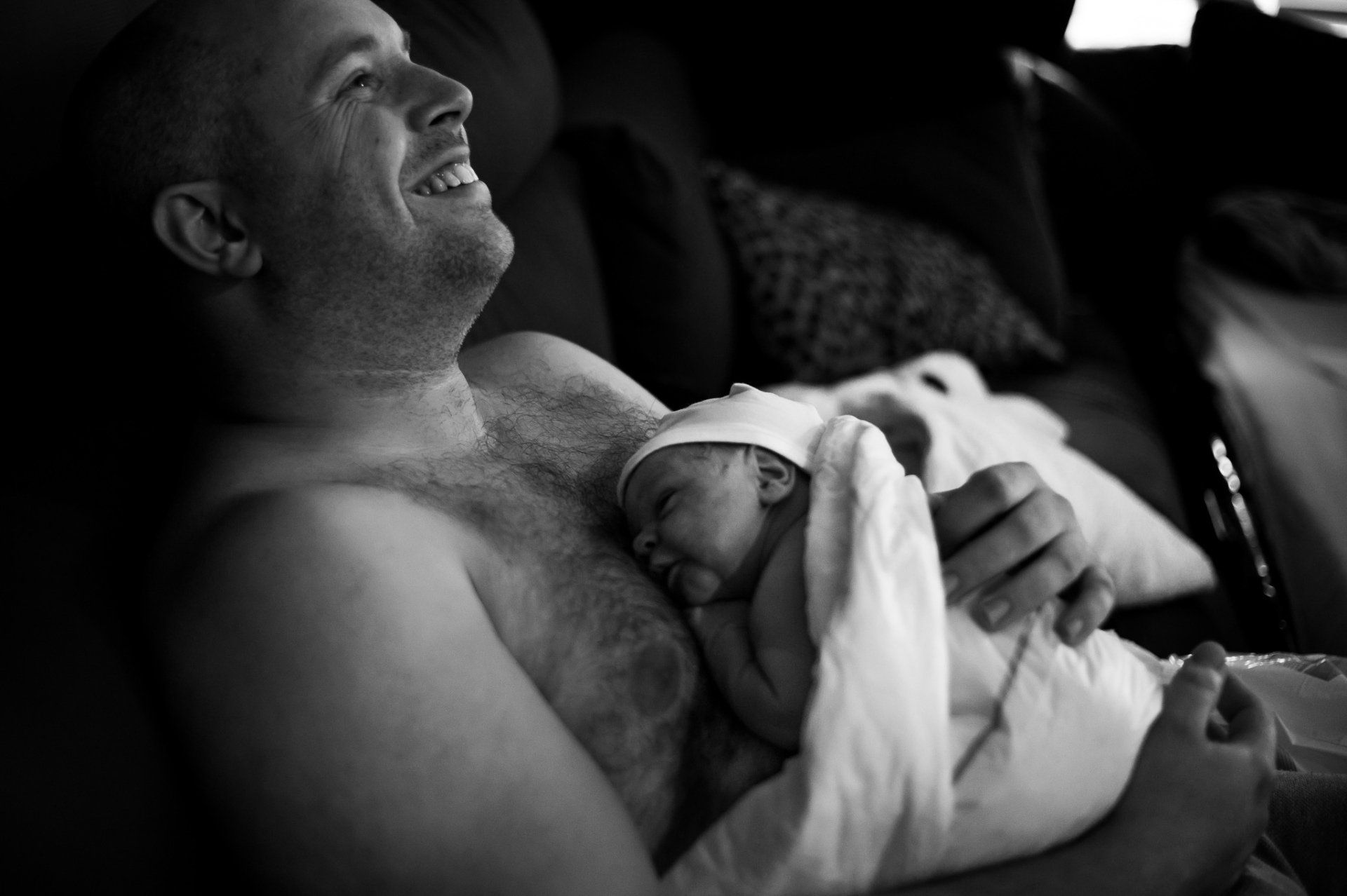 Een lachende man houdt een pasgeboren baby vast, gewikkeld in een deken. Zwart-witfoto.