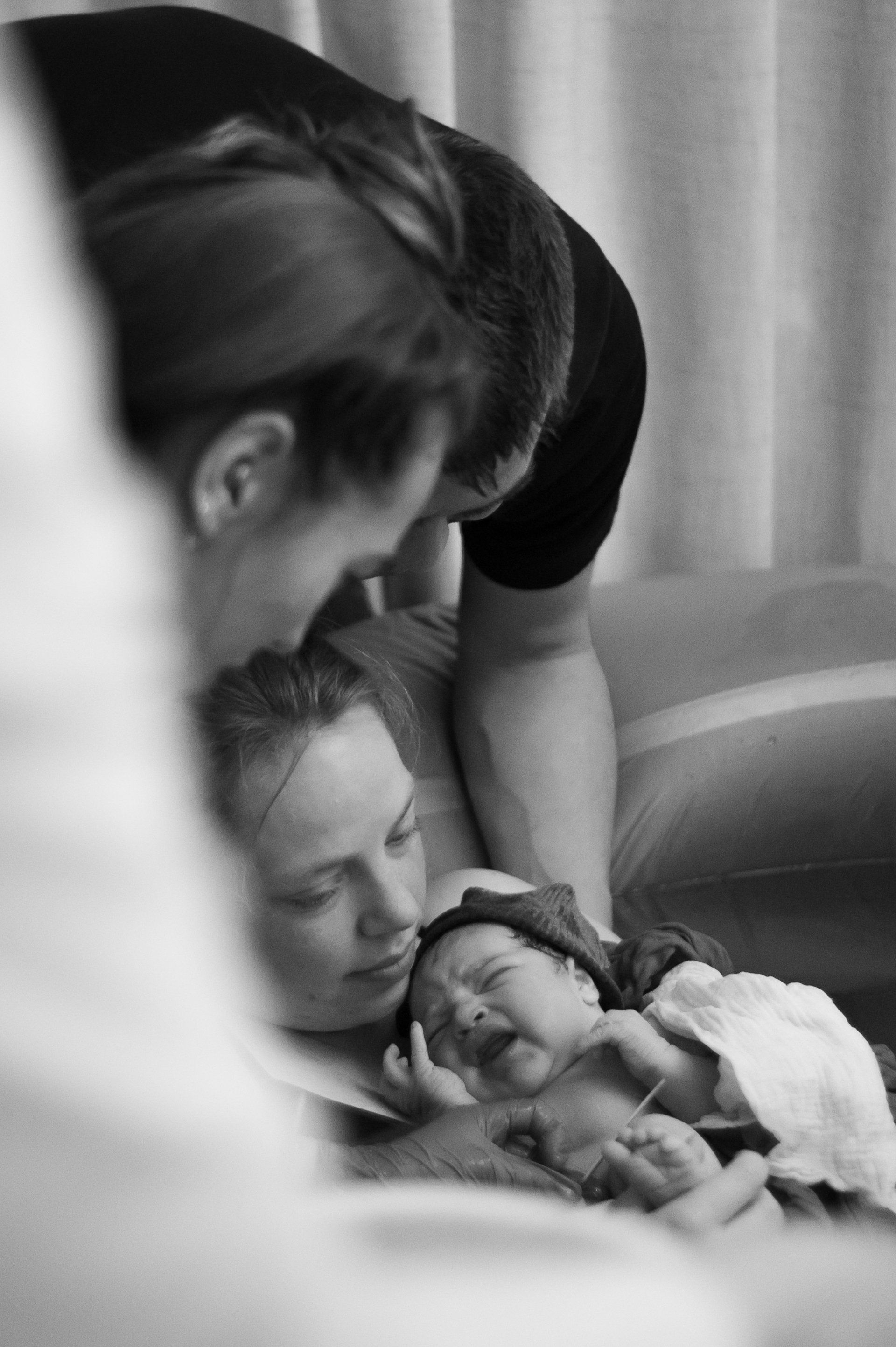 Pasgeboren baby in de armen van de moeder, vader buigt zich voorover om de moeder een kus op haar hoofd te geven. Zwart-wit, binnenopname.
