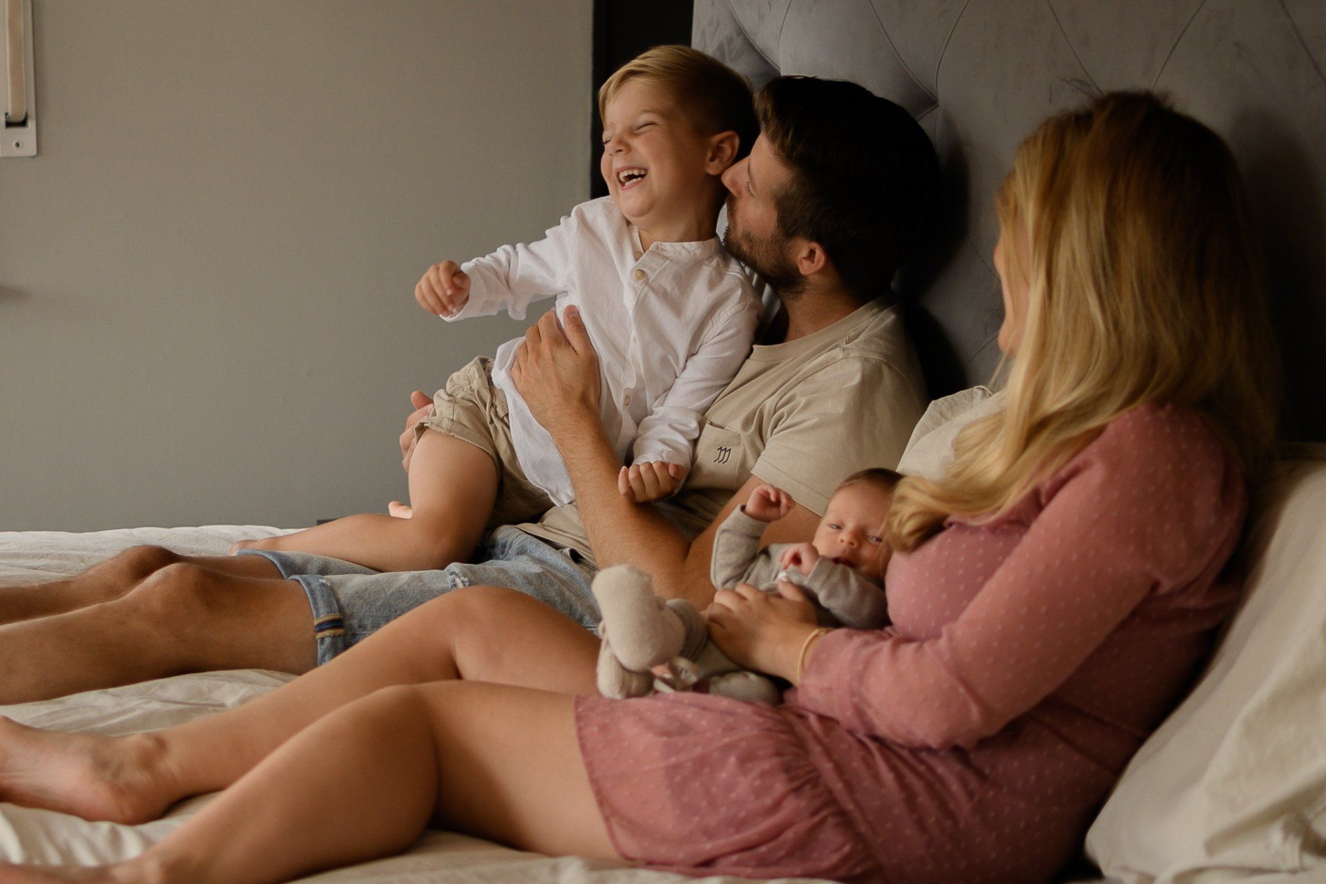 Een gezin van vier op een bed: vader met peuter, moeder met pasgeborene. Iedereen lacht.