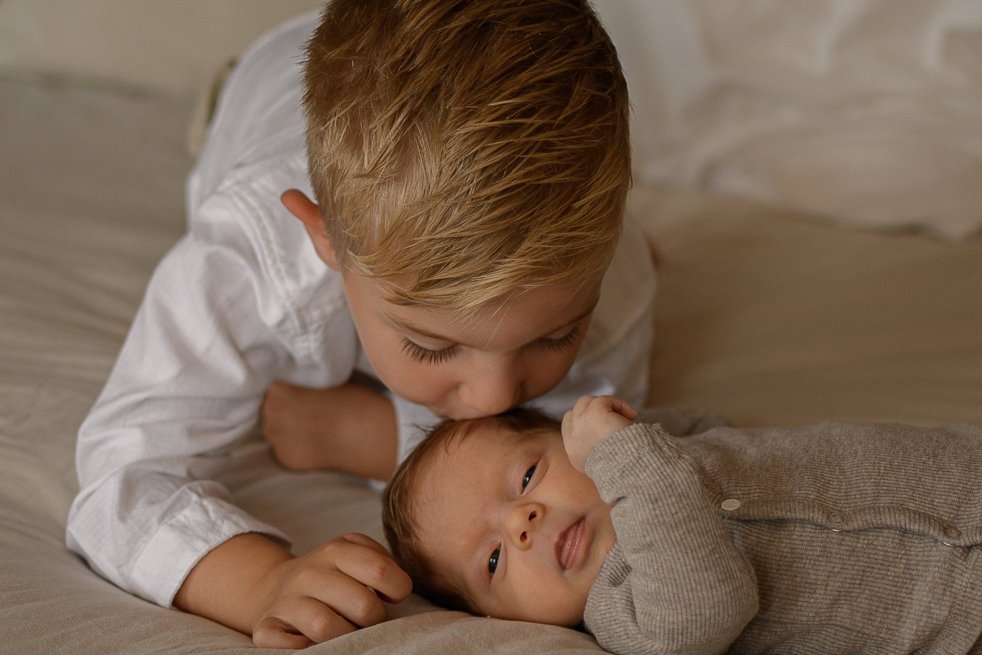 Een ouder kind kust een baby op het voorhoofd. Ze liggen allebei op een bed.
