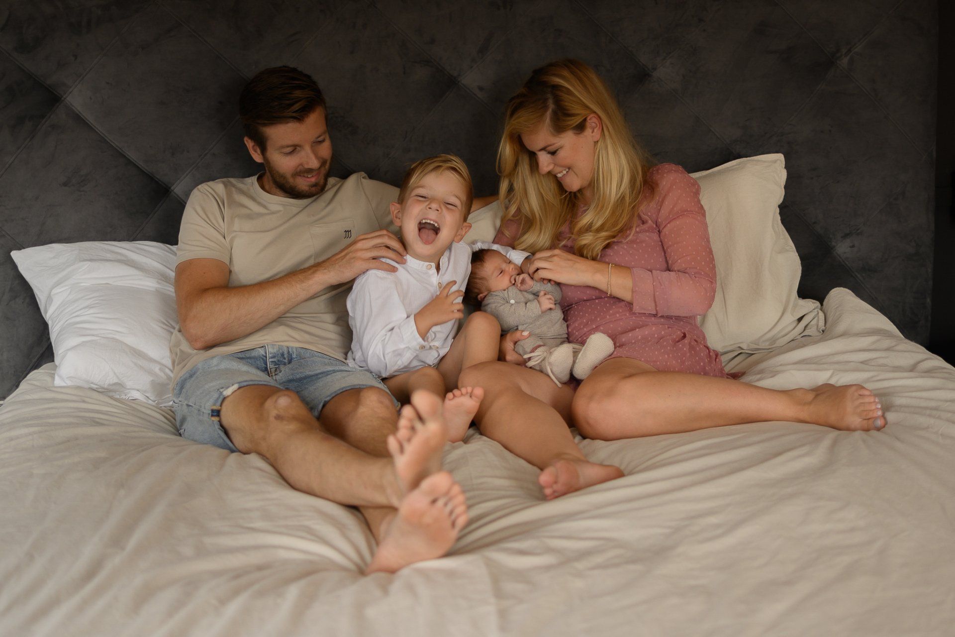 Een gezin van vier op een bed: ouders, een lachend ouder kind en een pasgeboren baby. Zachte verlichting, neutrale tinten.