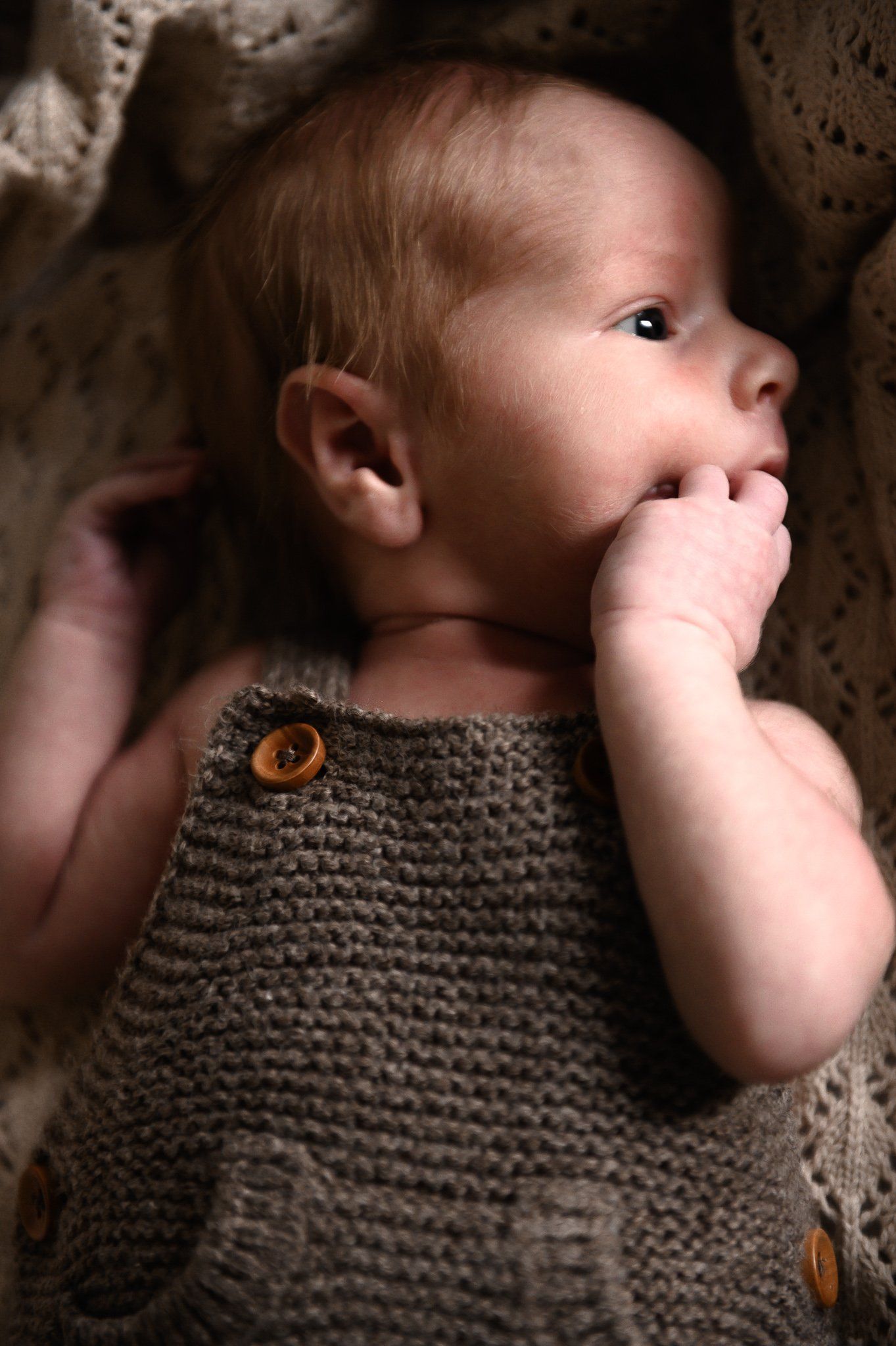 Pasgeboren baby in een gebreide bruine overall, liggend op een deken met patroon, kijkt naar rechts.