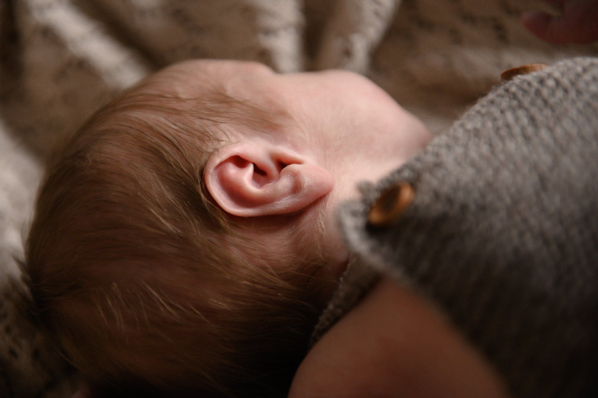 Close-up van het hoofdje van een baby, met een oortje zichtbaar, rustend op een dekentje en gekleed in een grijs gebreid kledingstuk.