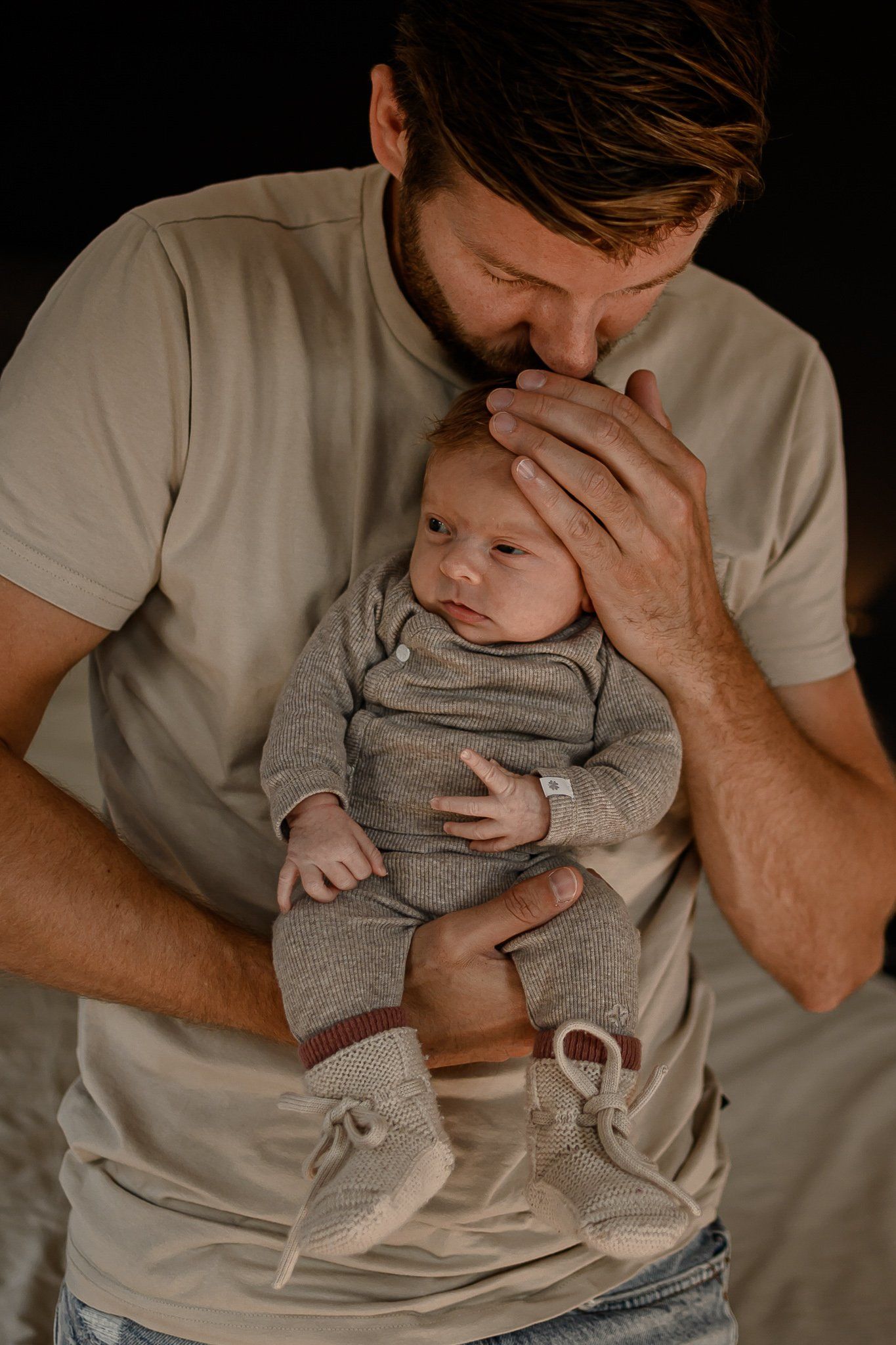Een man houdt het hoofdje van een baby vast en kust het. De baby draagt ​​een grijs pakje en een beige shirtje en bevindt zich binnen.