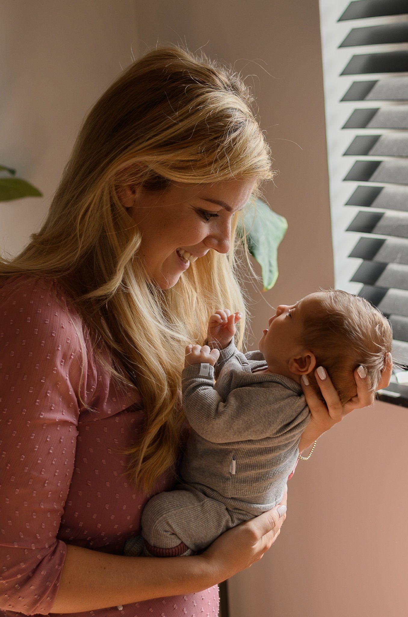 Een vrouw glimlacht terwijl ze een pasgeboren baby vasthoudt bij een raam met jaloezieën. Het zonlicht stroomt naar binnen.