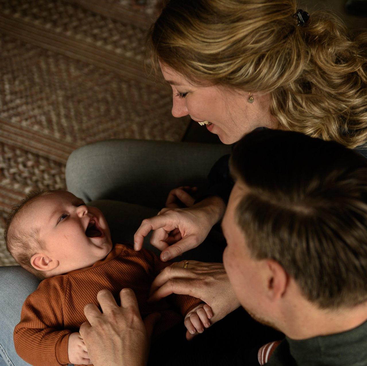 Ouders kietelen een baby die op schoot ligt, en ze lachen allemaal vrolijk.