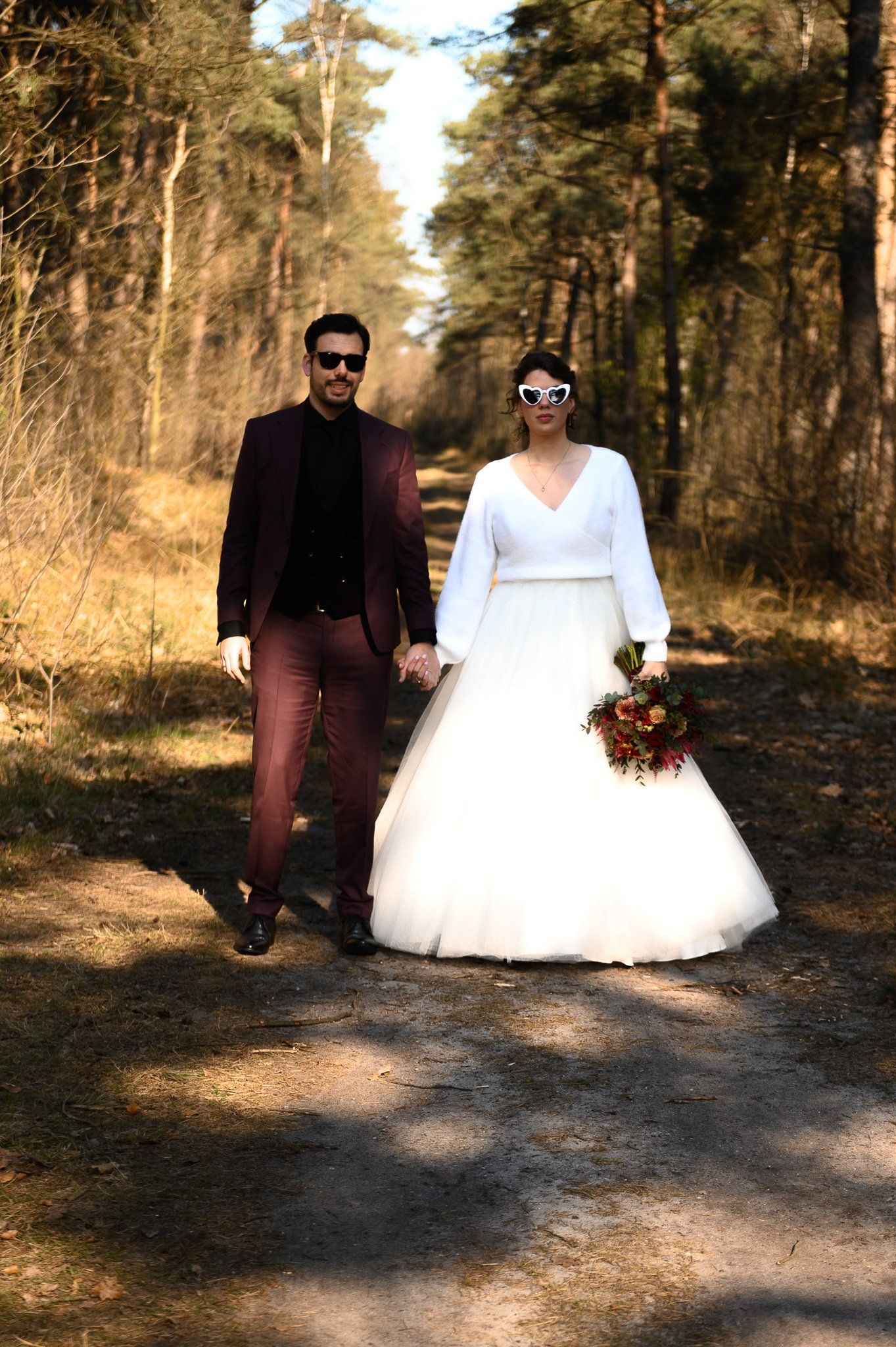 Een echtpaar in trouwkleding loopt hand in hand over een pad in het bos; de man draagt ​​een bordeauxrood pak, de vrouw een witte jurk.
