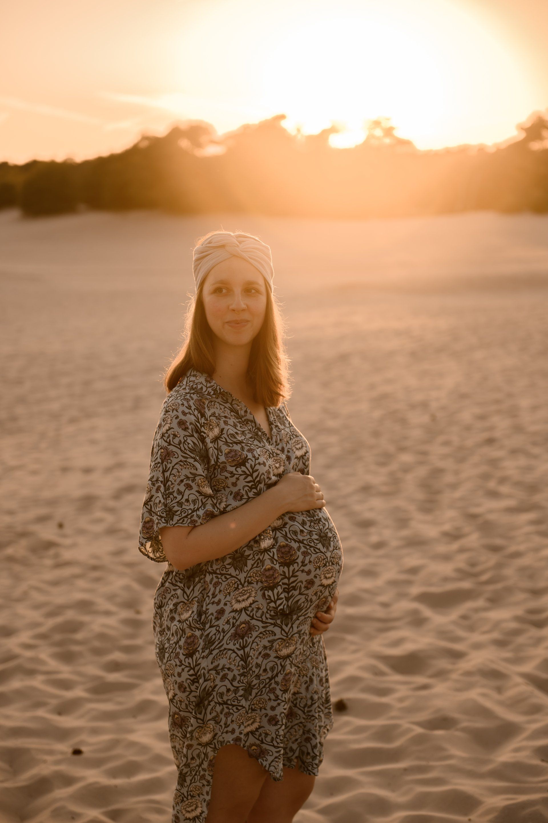 Een zwangere vrouw in een jurk met patroon staat op een zandstrand en houdt haar buik vast, met de zonsondergang op de achtergrond.