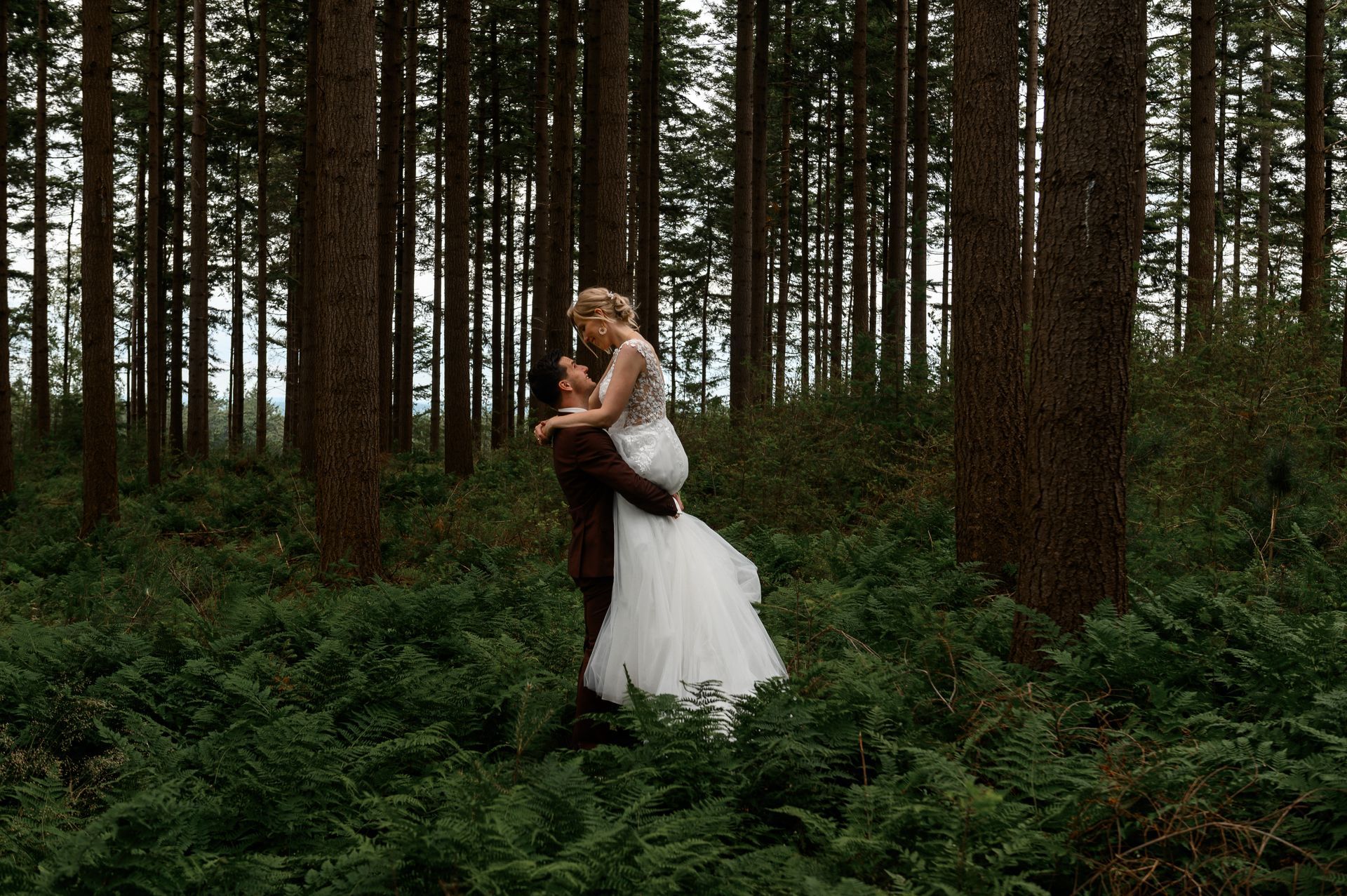 Een stel omhelst elkaar in een bos. De man tilt de vrouw in een witte trouwjurk op te midden van groene varens en hoge bomen.