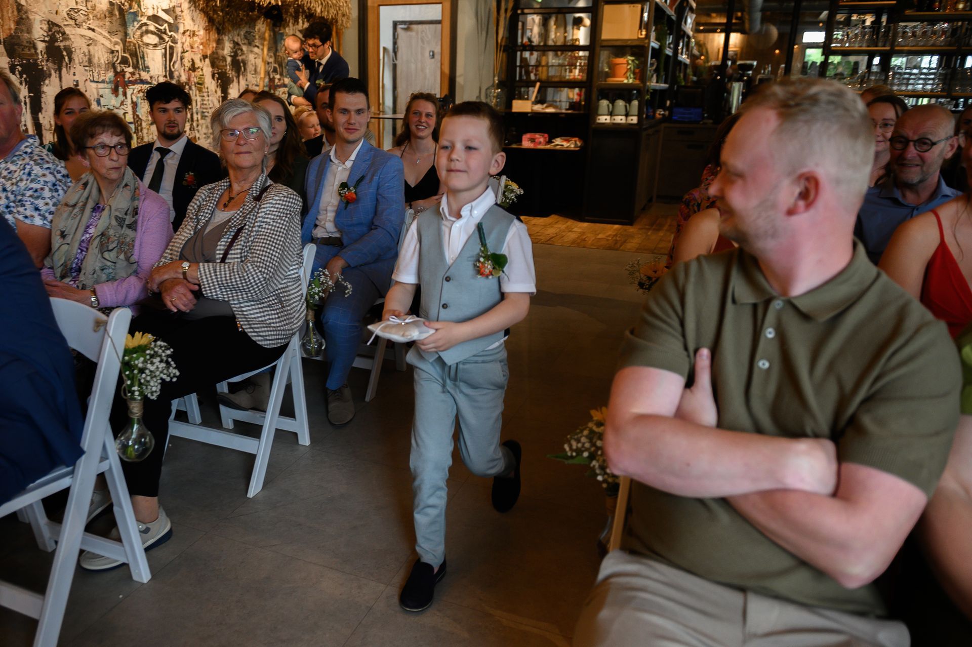 Een jonge jongen in een grijs pak loopt met een dienblad door het gangpad tijdens een ceremonie. Gasten zitten aan beide zijden.