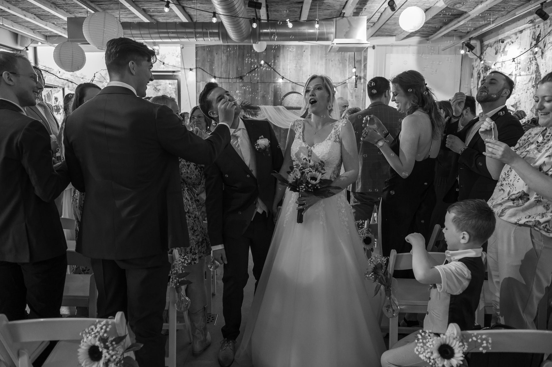 Huwelijksceremonie: Bruid en bruidegroom omringd door gasten, lachend, confetti, binnenlocatie.