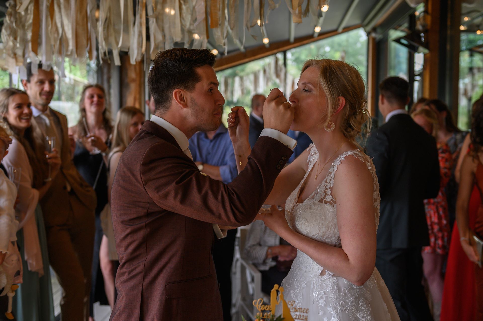 Bruid en bruidegom voeren elkaar taart tijdens een huwelijksreceptie. Ze glimlachen en worden omringd door gasten.