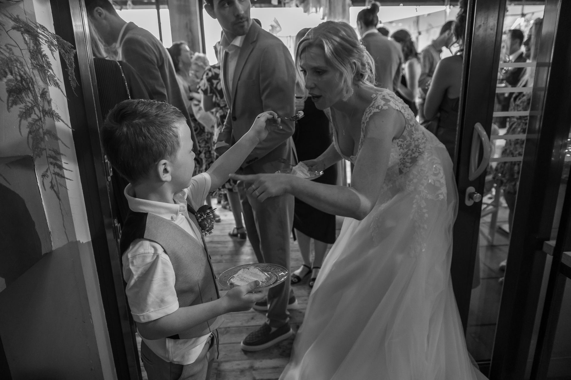 Bruid en jonge jongen delen taart op een bruiloft. Vrouw in trouwjurk geeft jongen te eten terwijl gasten toekijken.