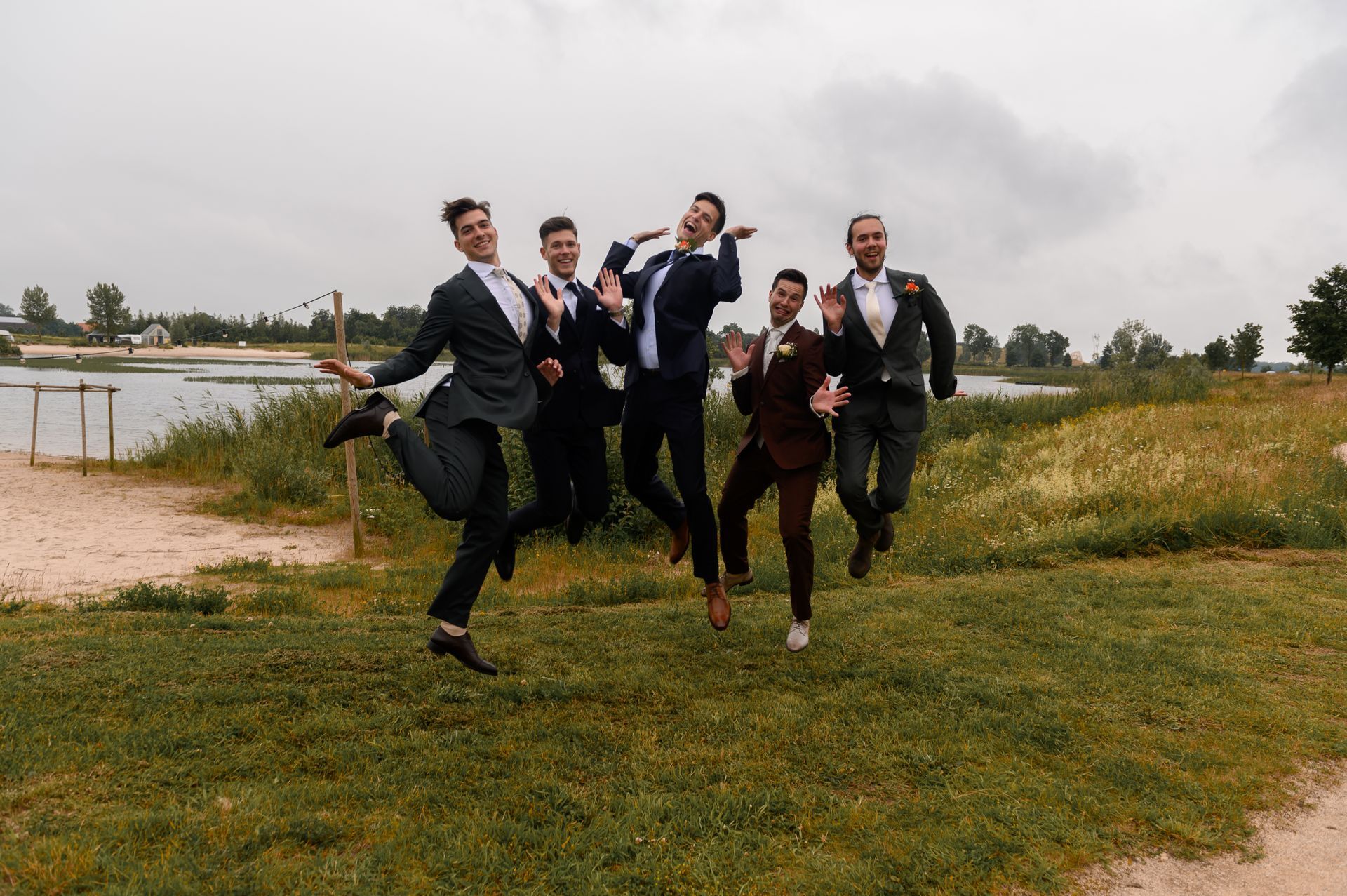 Mannen in pakken springen van vreugde bij een meer. Groen gras, bewolkte lucht.