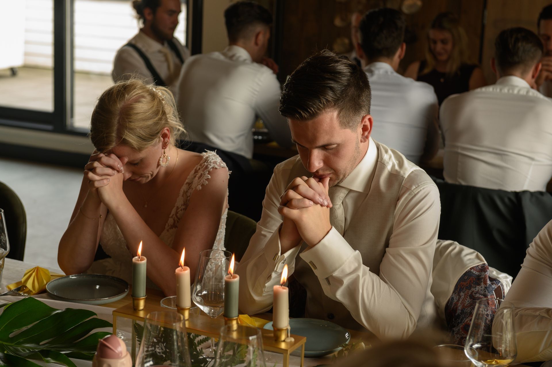 Bruid en bruidegom op een bruiloft, hoofden gebogen in gebed, handen ineengevouwen. Kaarsen op tafel.