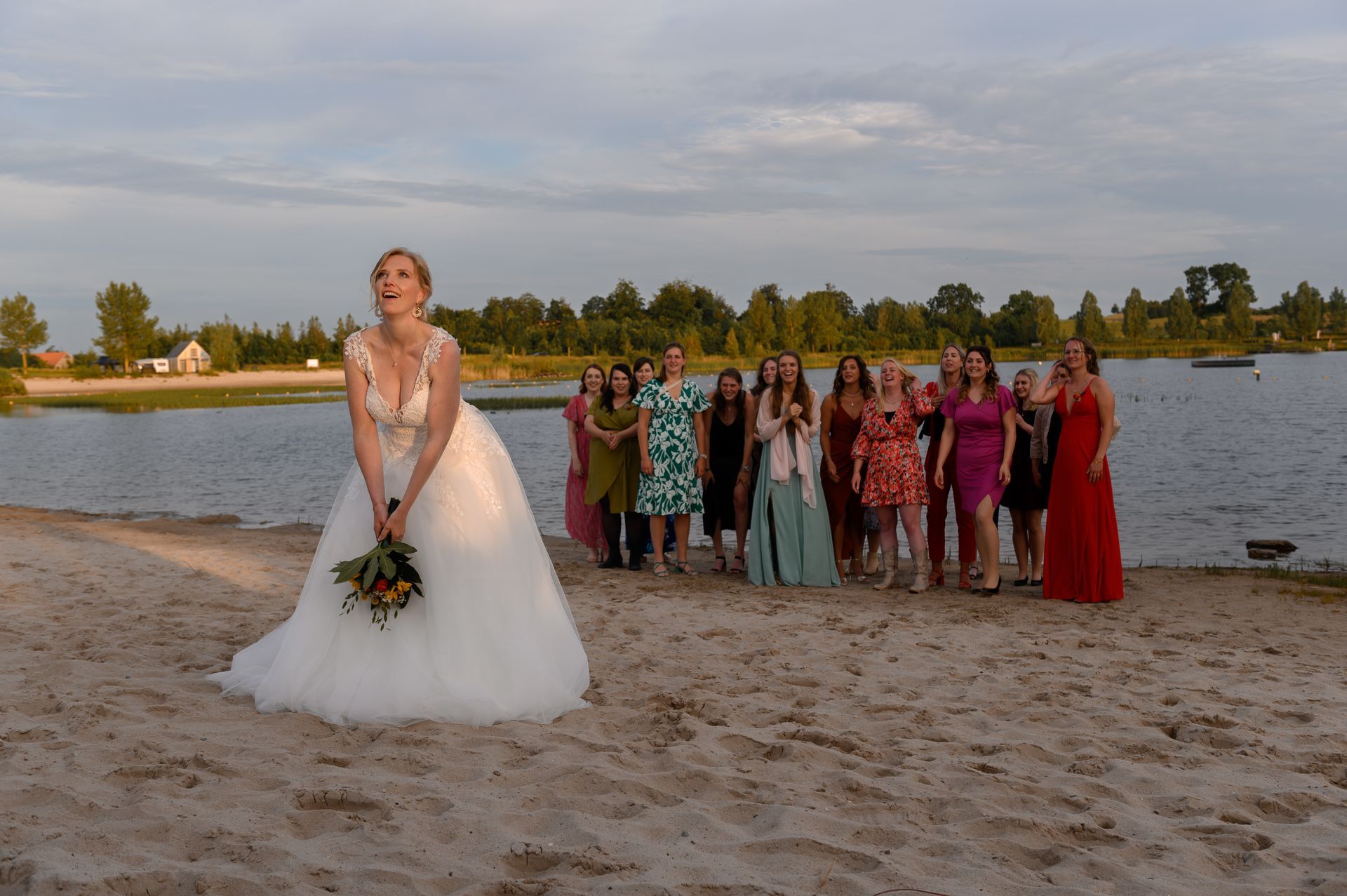Bruid gooit boeket naar groep vrouwen op het strand. Meer en bomen op de achtergrond.