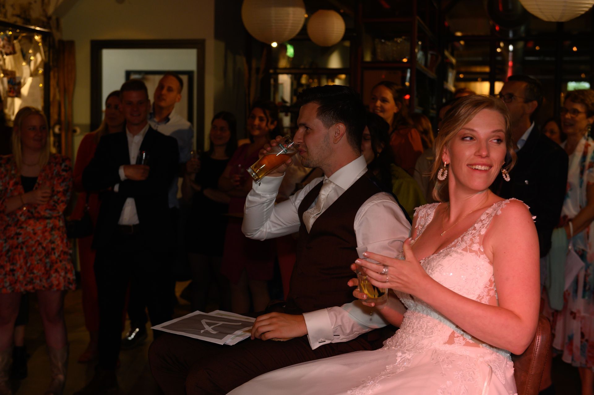 Pasgetrouwden op een receptie, de bruidegom drinkt, de bruid glimlacht. Gasten kijken toe, verlicht door warm licht.