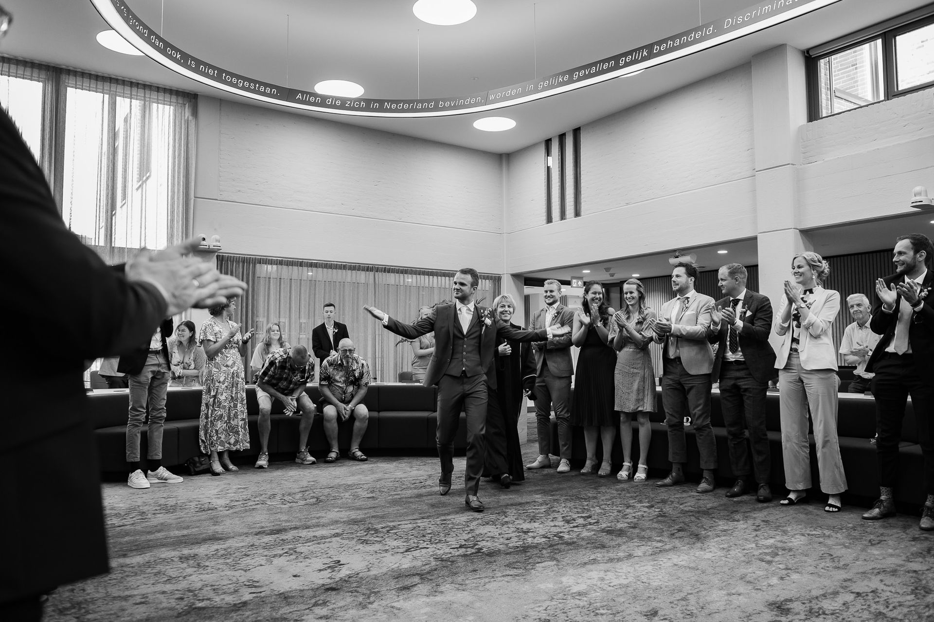 De bruidegom verlaat de ceremonie met opgeheven arm en wordt begroet door applaudisserende gasten. Moderne interieursetting.