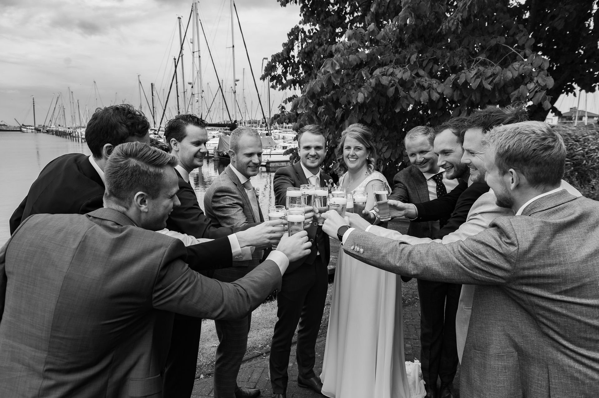 Het bruidspaar en de gasten proosten met champagne; bruid en bruidegroom in het midden. Boten in de haven.
