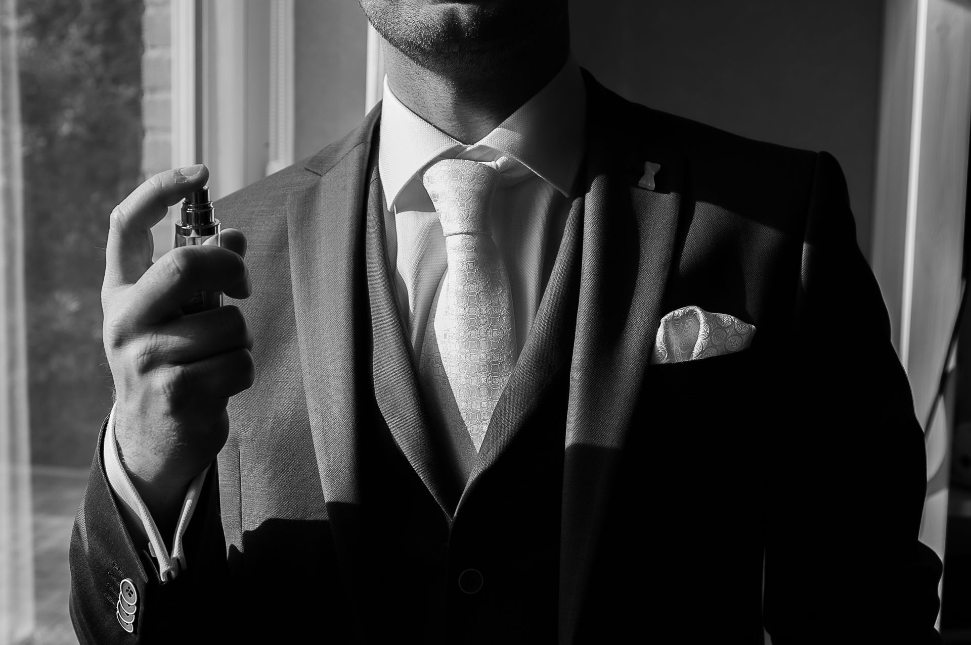 Man in pak spuit parfum bij een raam, zwart-wit.