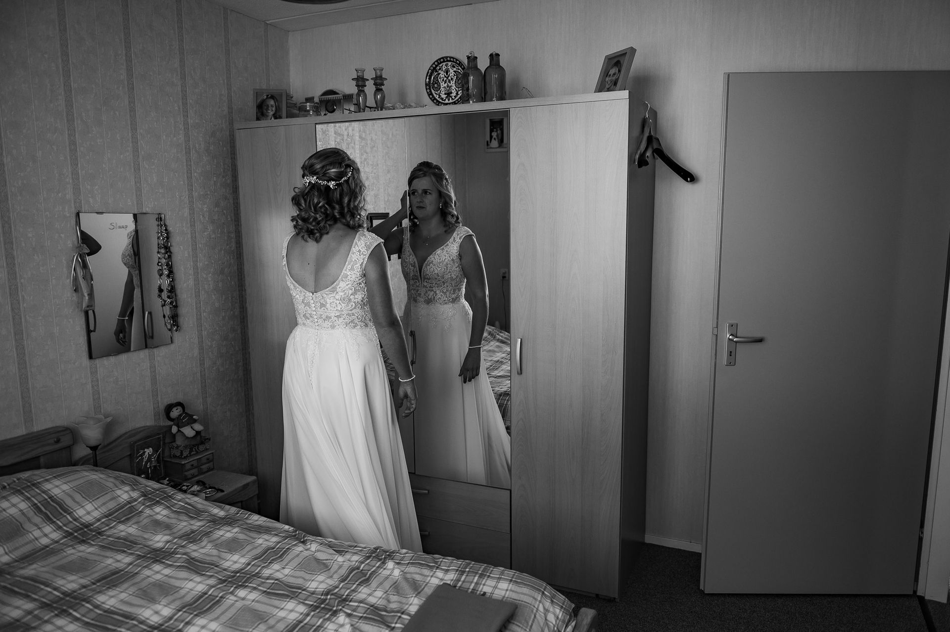 Bruid in trouwjurk die naar haar spiegelbeeld kijkt in een grote spiegel in een kledingkast in een slaapkamer.