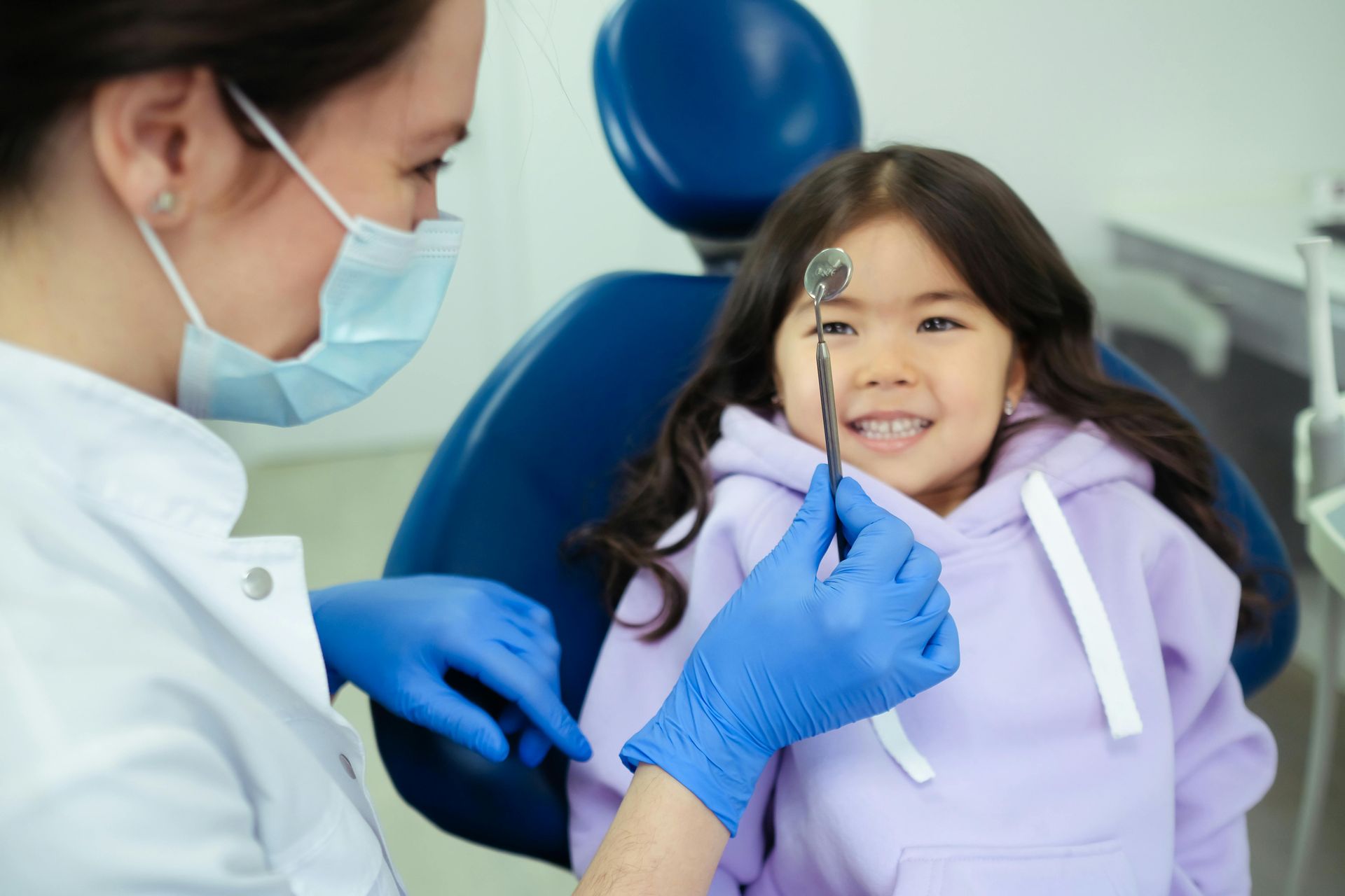 Dentist examining a smiling child’s teeth in a dental chair, wearing blue gloves and a mask